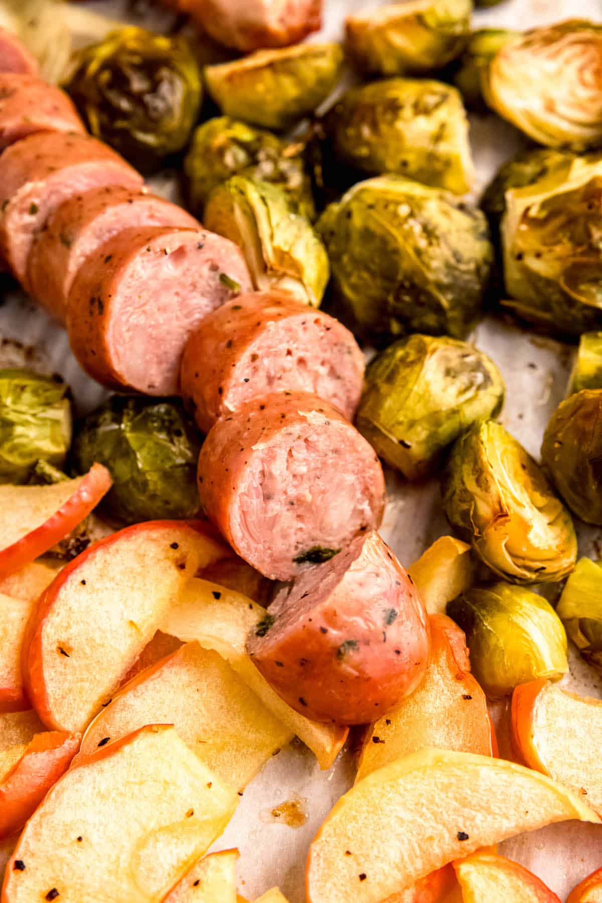 closeup shot of sliced chicken sausage on a sheet pan with roasted brussels sprouts and apples.