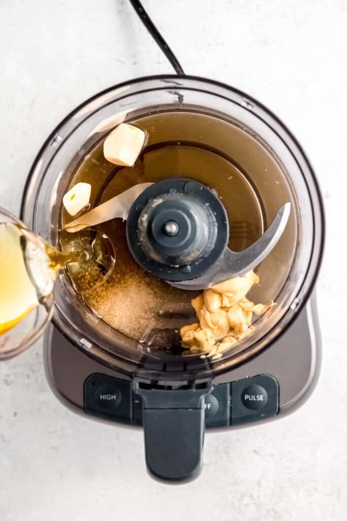 action shot of pouring oil into the base of a food processor with the remaining creamy maple dijon apple cider vinaigrette ingredients.