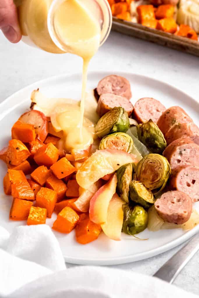 action shot of a hand drizzling creamy maple dijon vinaigrette over a plate of sheet pan chicken sausage with brusssels sprouts, apples, onions, fennel, and sweet potato.