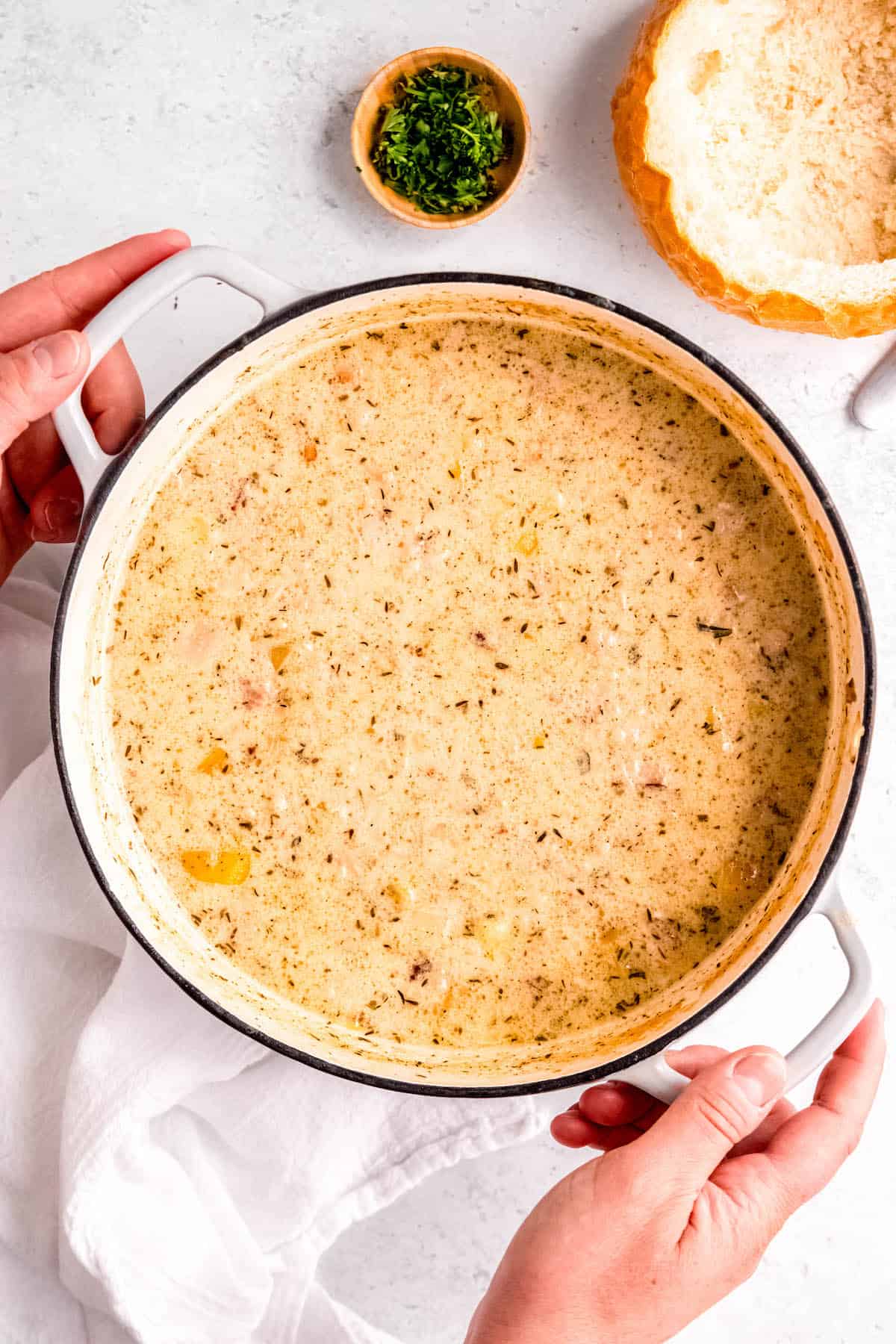 hands grabbing the handles of the white dutch oven filled with homemade gluten-free clam chowder.