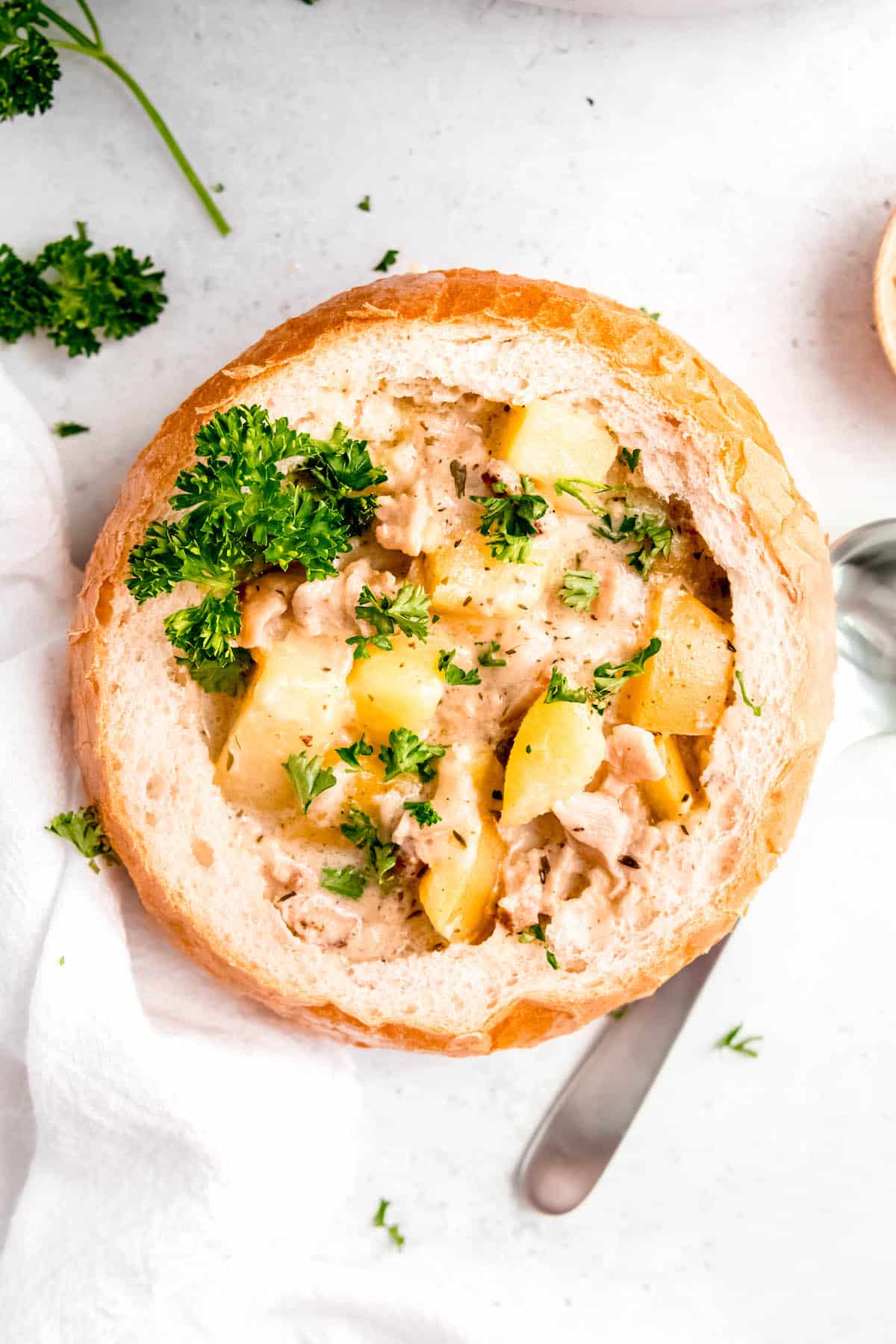 bread bowl filled with clam chowder on a white table with a silver spoon and parsley scattered around.