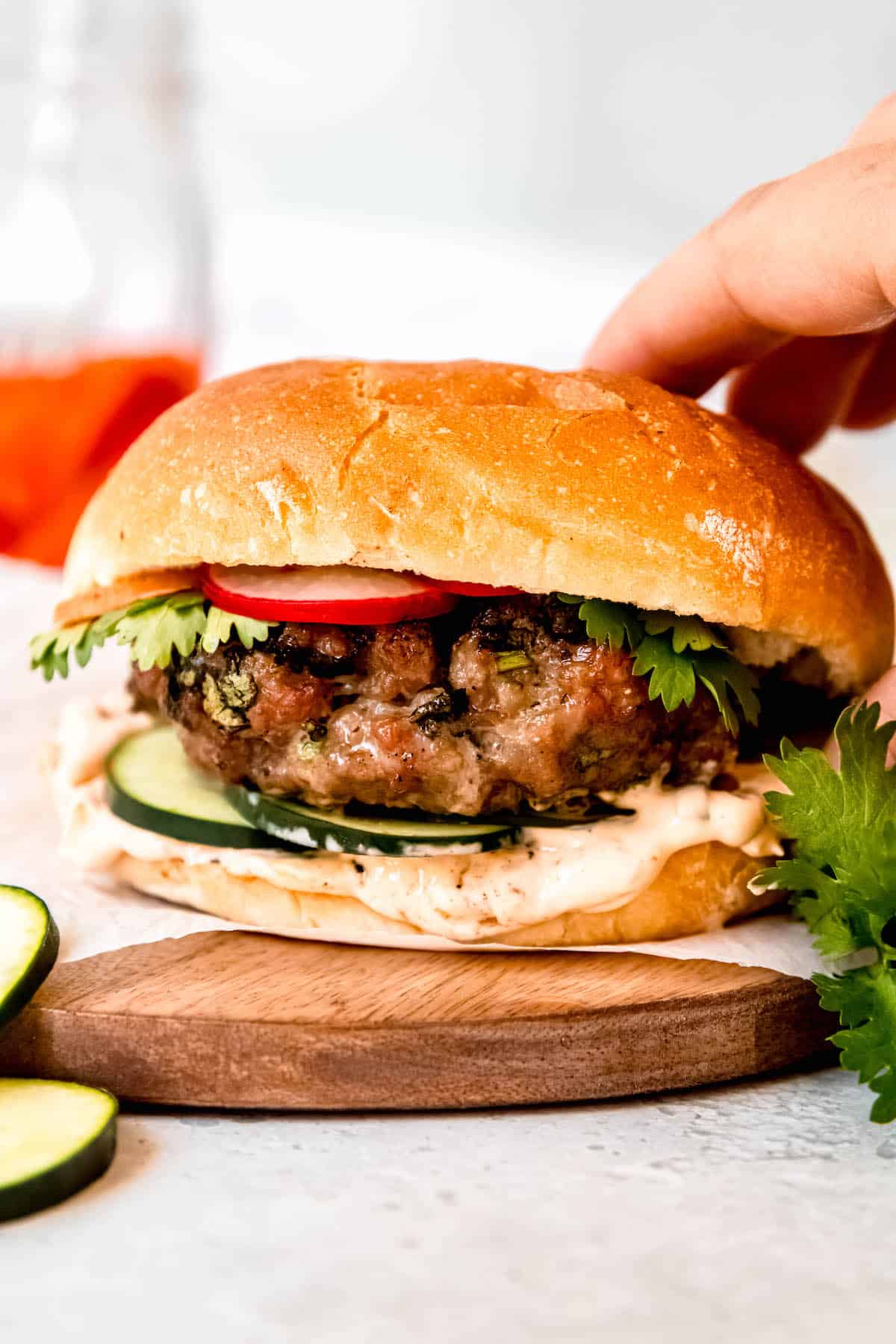 hand reaching out to grab a homemade Asian pork burger with sriracha mayo and pickled banh mi vegetables from a wooden serving board.