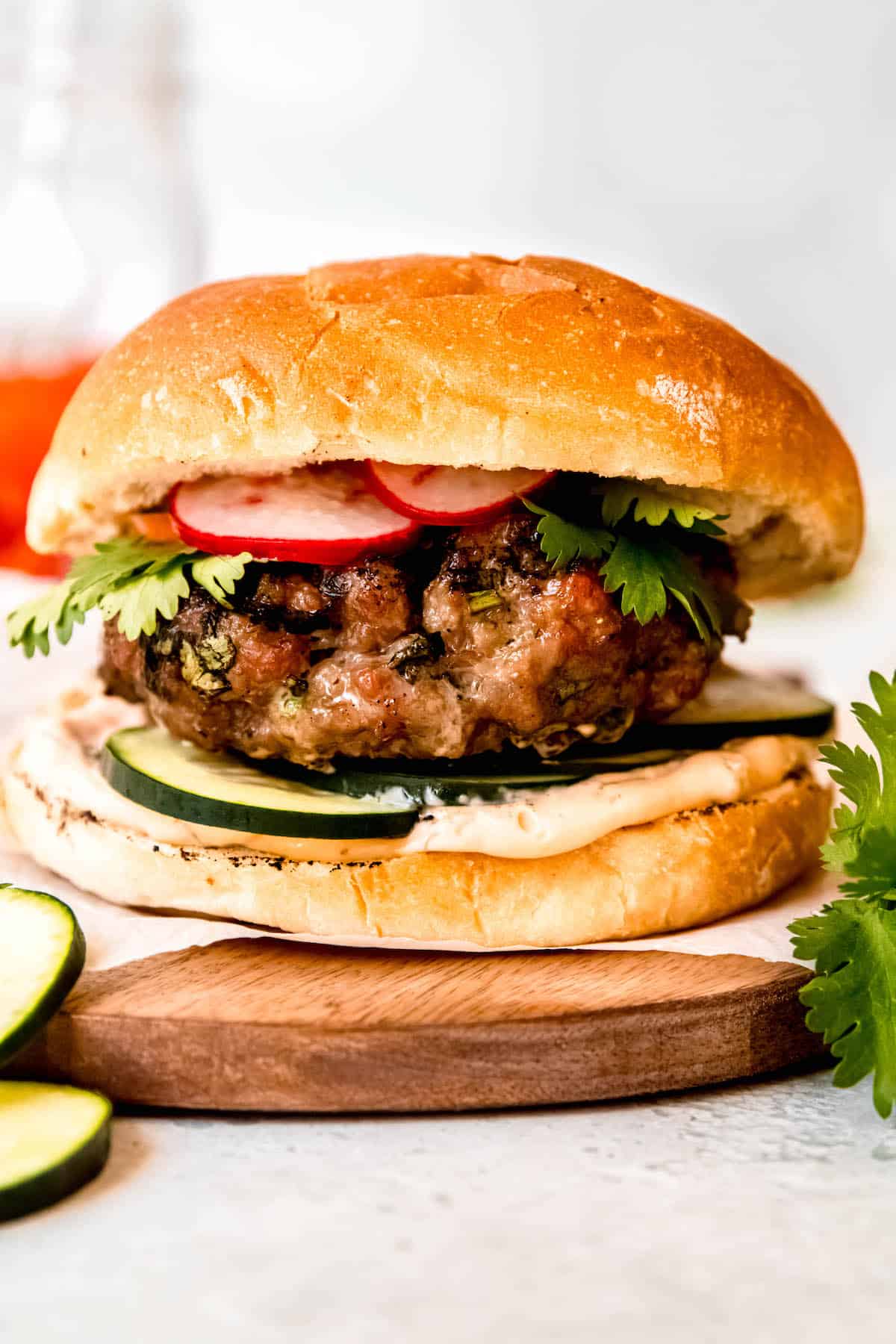 side-on view of an Asian pork burger showing the toasted bun, spicy mayo, fresh cucumber slices, green onion-flecked grilled pork patty, pickled banh mi veggies, and fresh cilantro all layered together.