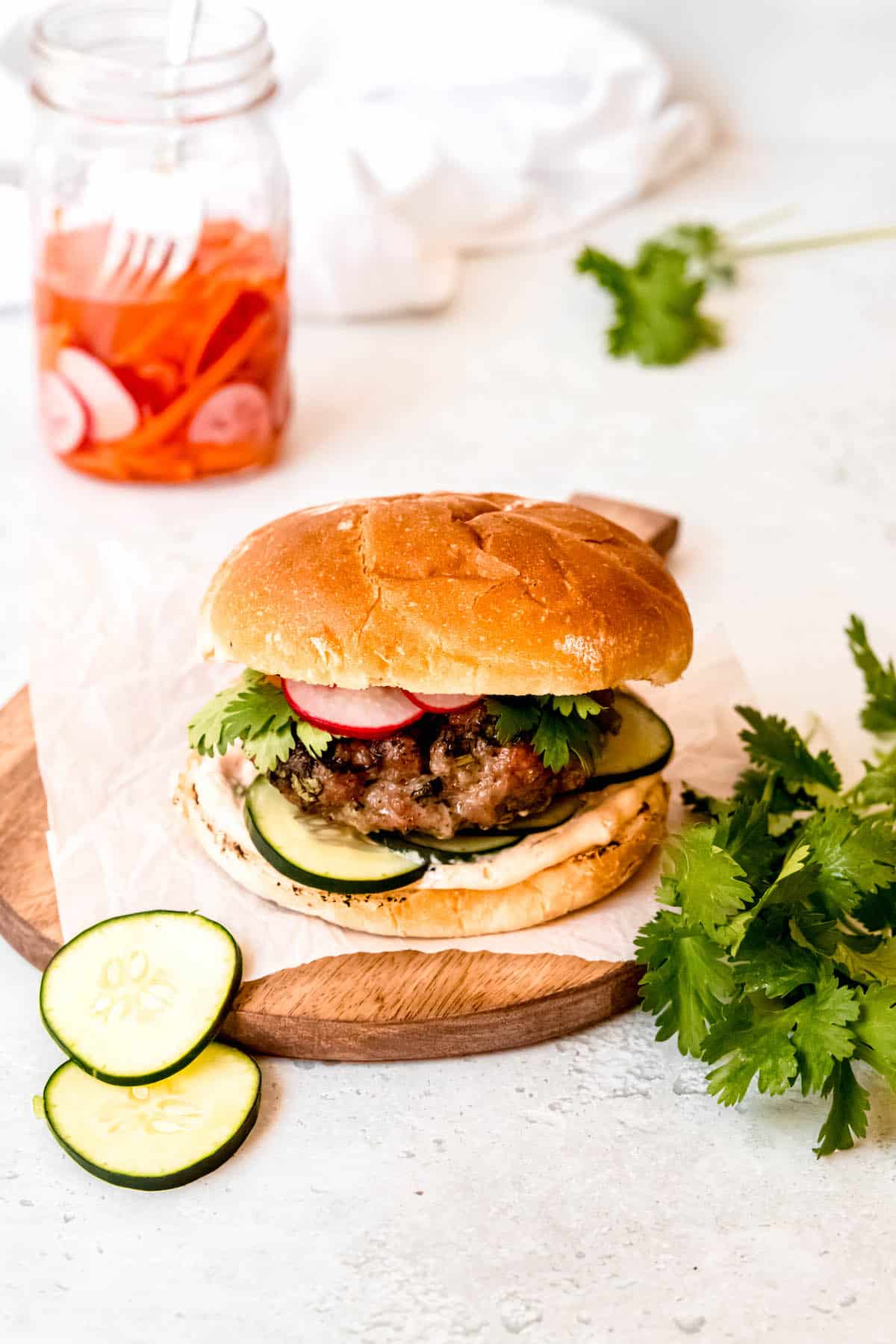 table set with a wooden serving board holding an Asian pork burger with a jar of pickled banh mi veggies, a bunch of fresh cilantro, and slices of fresh cucumber around.