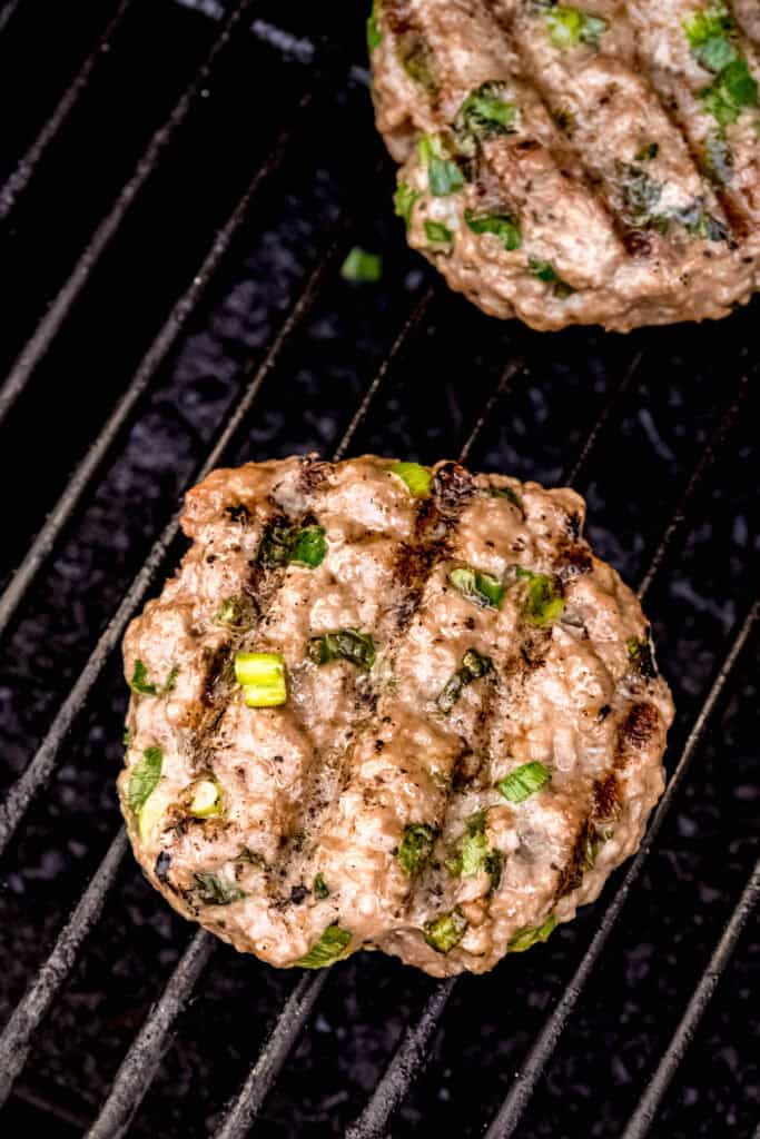 Asian-flavored pork burger patties with green onions and fish sauce on the grates of a grill; you can see the char marks from grilling the first side.