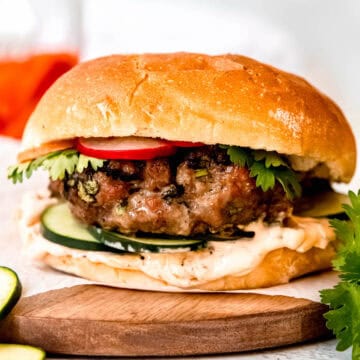 juicy Asian pork burger on a piece of parchment paper on a round wooden tray with a vibrant jar of pickled carrots and radishes in the background and fresh cucumber and cilantro in the foreground.