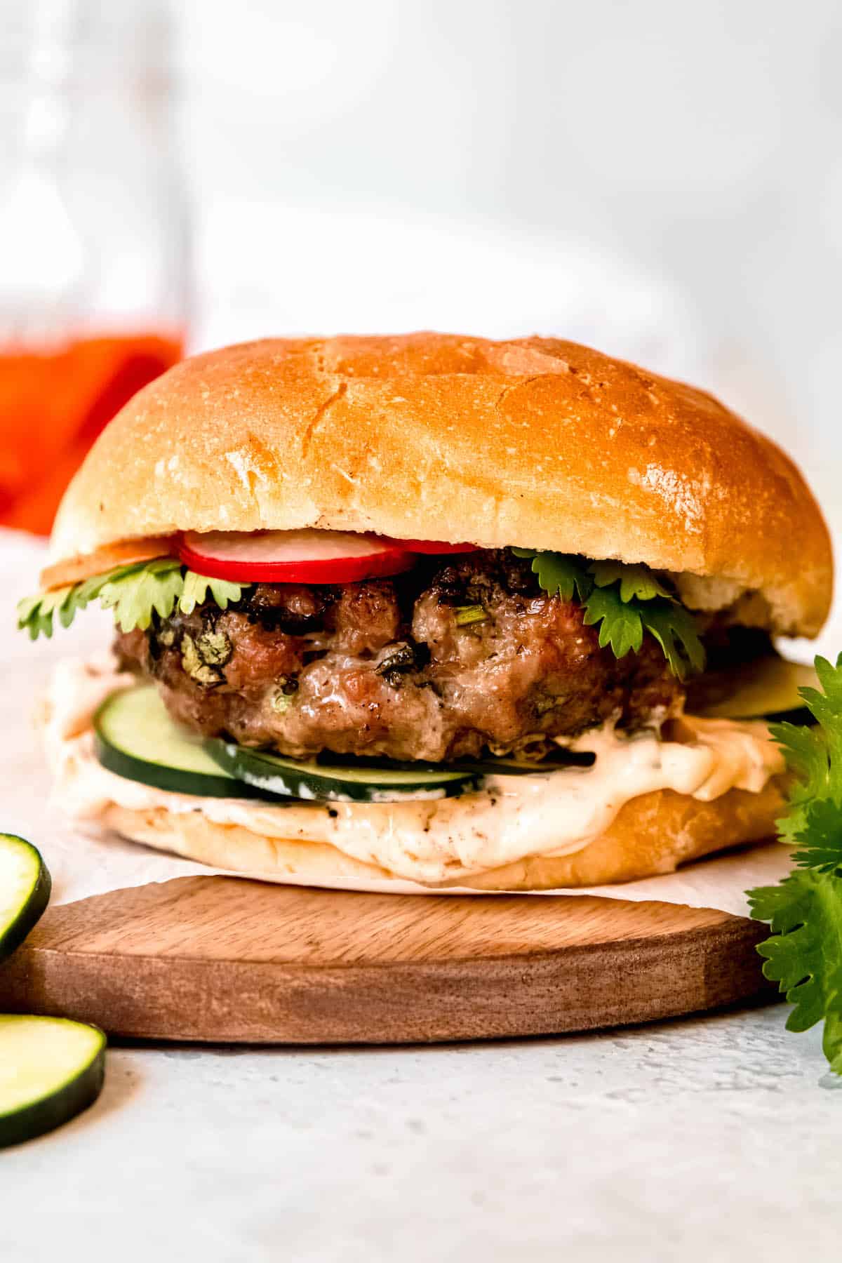 juicy Asian pork burger on a piece of parchment paper on a round wooden tray with a vibrant jar of pickled carrots and radishes in the background and fresh cucumber and cilantro in the foreground.