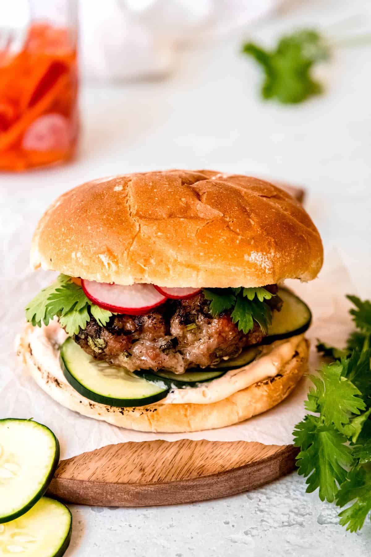 45-degree angle shot of an Asian-inspired banh mi pork burger on a piece of parchment atop a wooden round on a white table with a bunch of fresh cilantro, fresh cucumber slices, and a jar of pickled carrots and radishes.