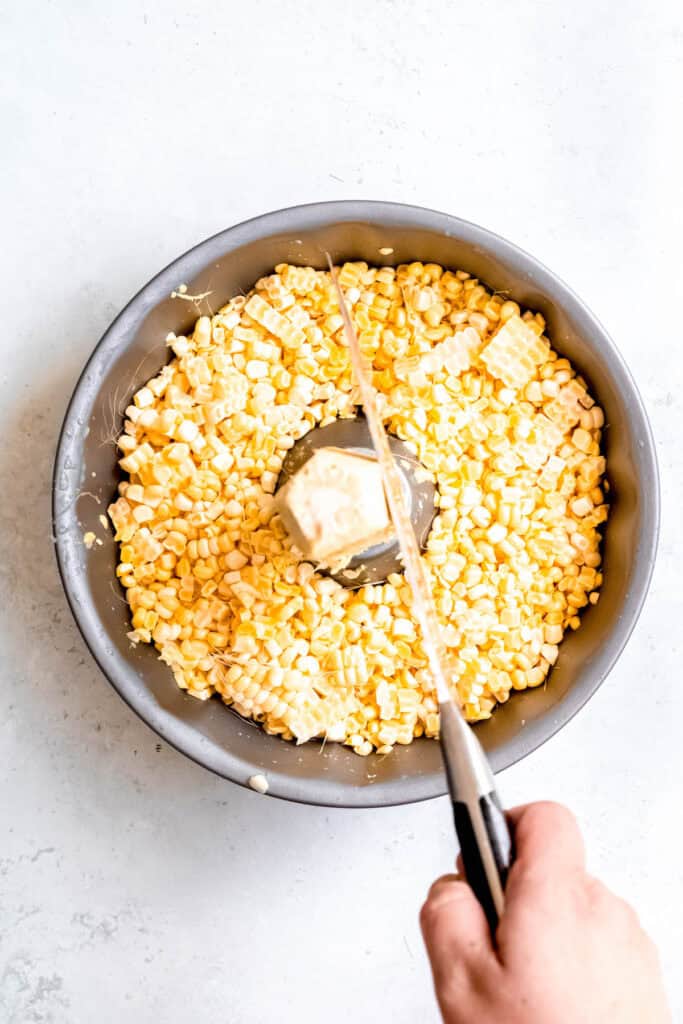 overhead shot of a hand holding a knife and slicing the kernels off a cob of corn that's standing upright in the center of a bundt pan with corn kernels filling the cake well.