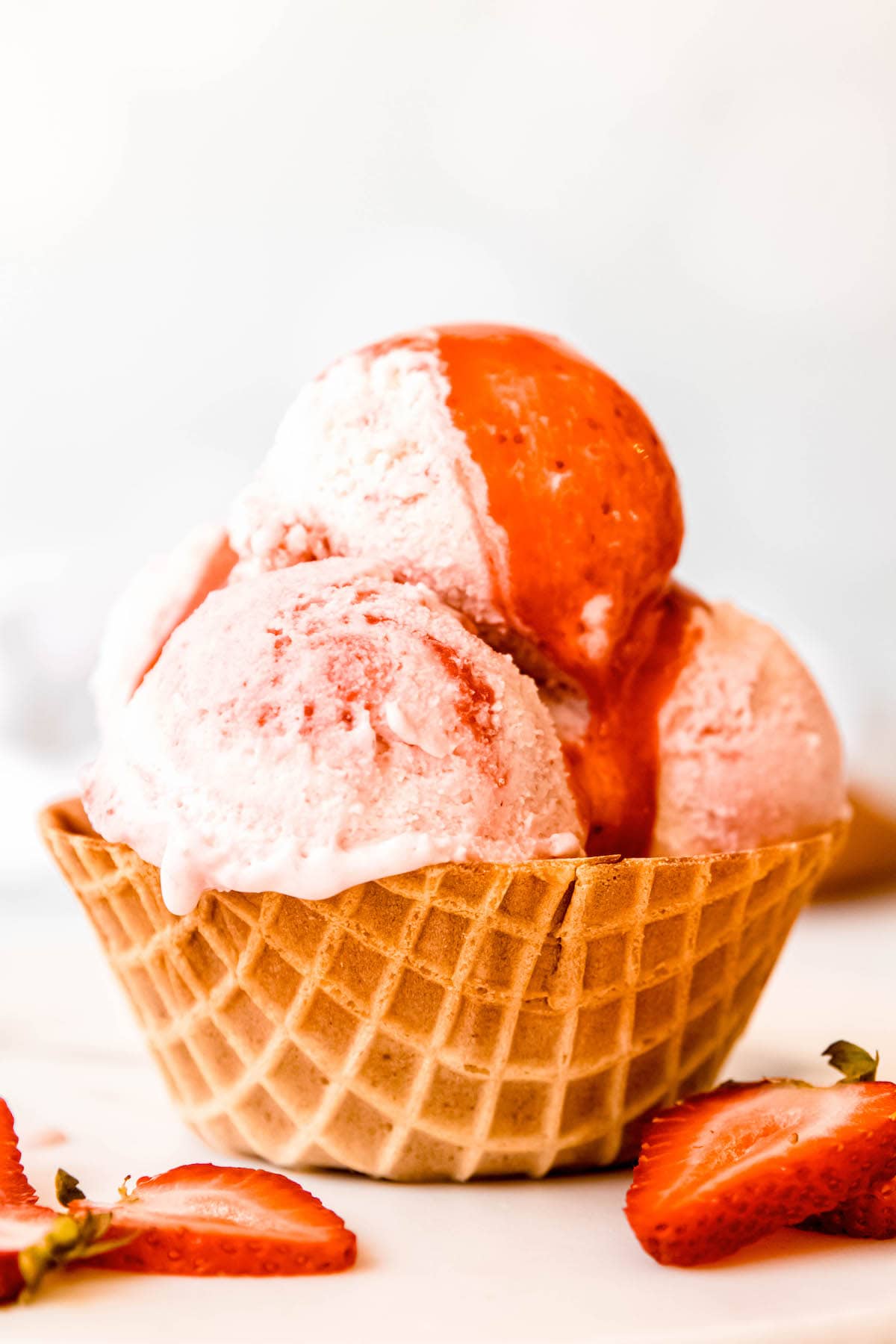 closeup shot of 3 scoops of strawberry swirl ice cream in a waffle cup topped with extra strawberry sauce and surrounded by fresh strawberry slices.