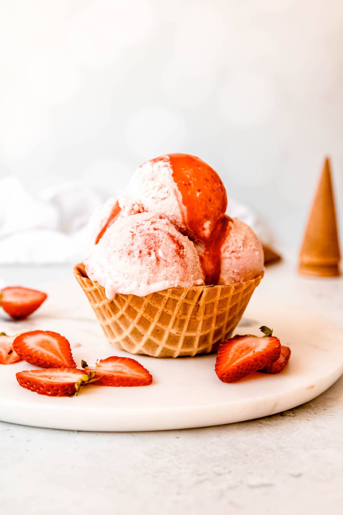 waffle bowl with 3 scoops of strawberry swirl ice cream on a white table with sliced strawberries and a stack of ice cream cones blurred out in the background.