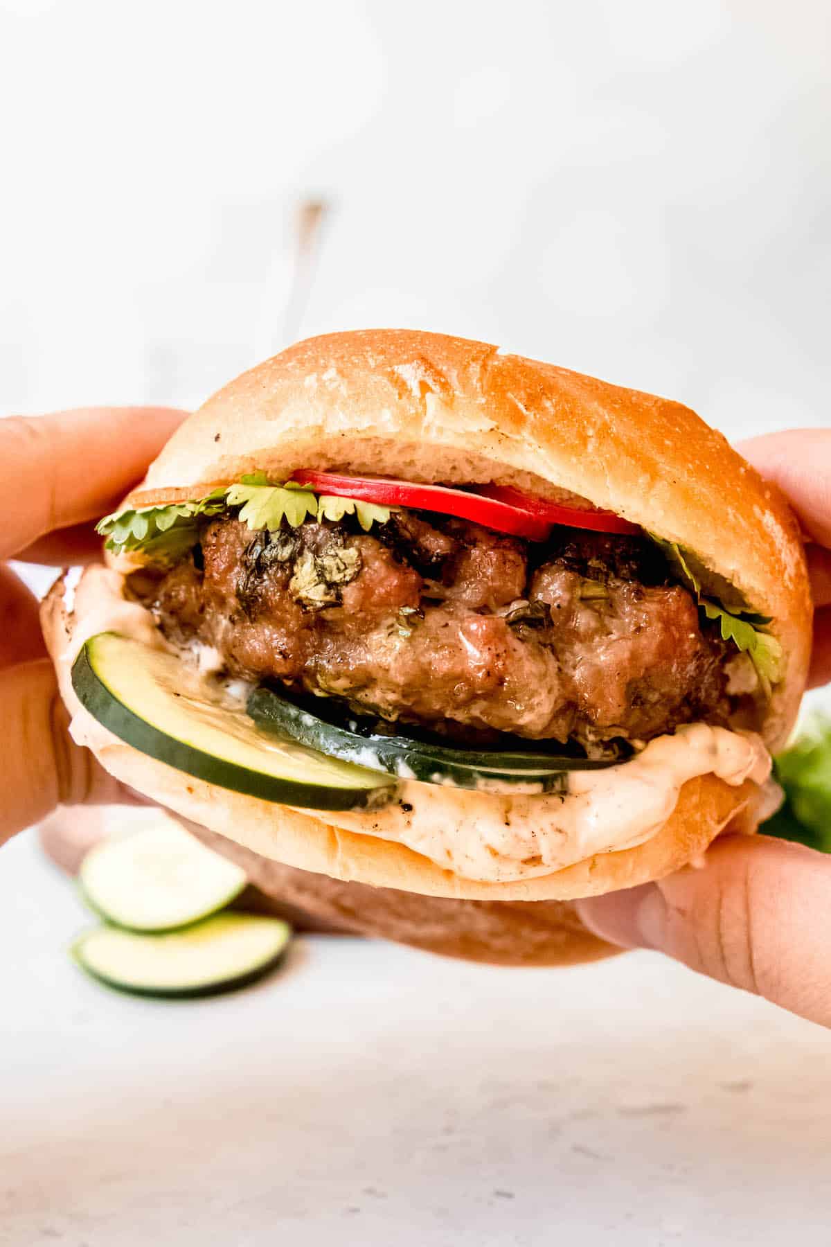 hands holding an Asian pork burger on a soft brioche bun loaded with pickled carrots and radishes, fresh cucumber and cilantro, and spicy mayo.