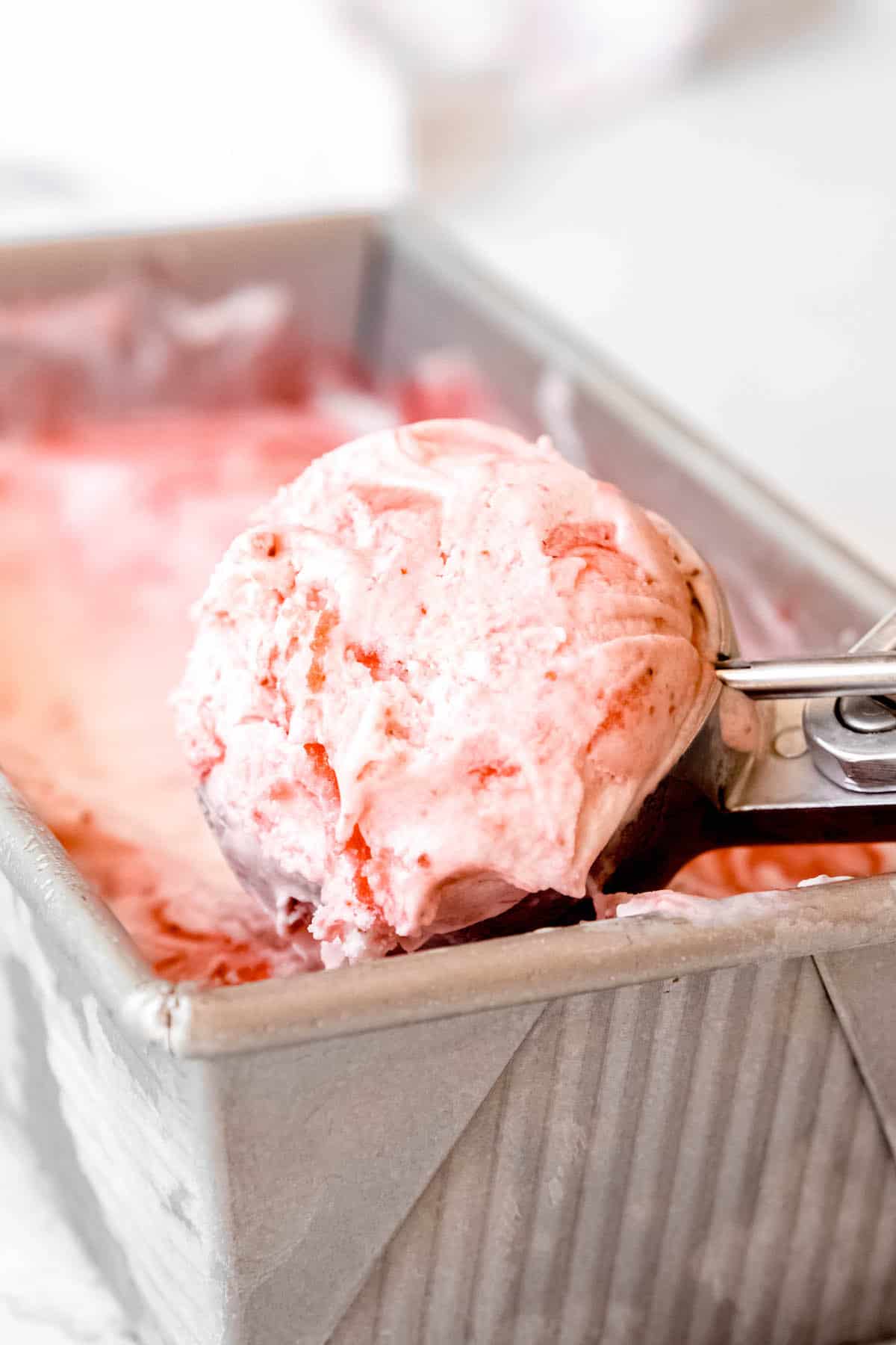 silver loaf tin with homemade strawberry swirl ice cream and an ice cream scoop taking a scoop out.
