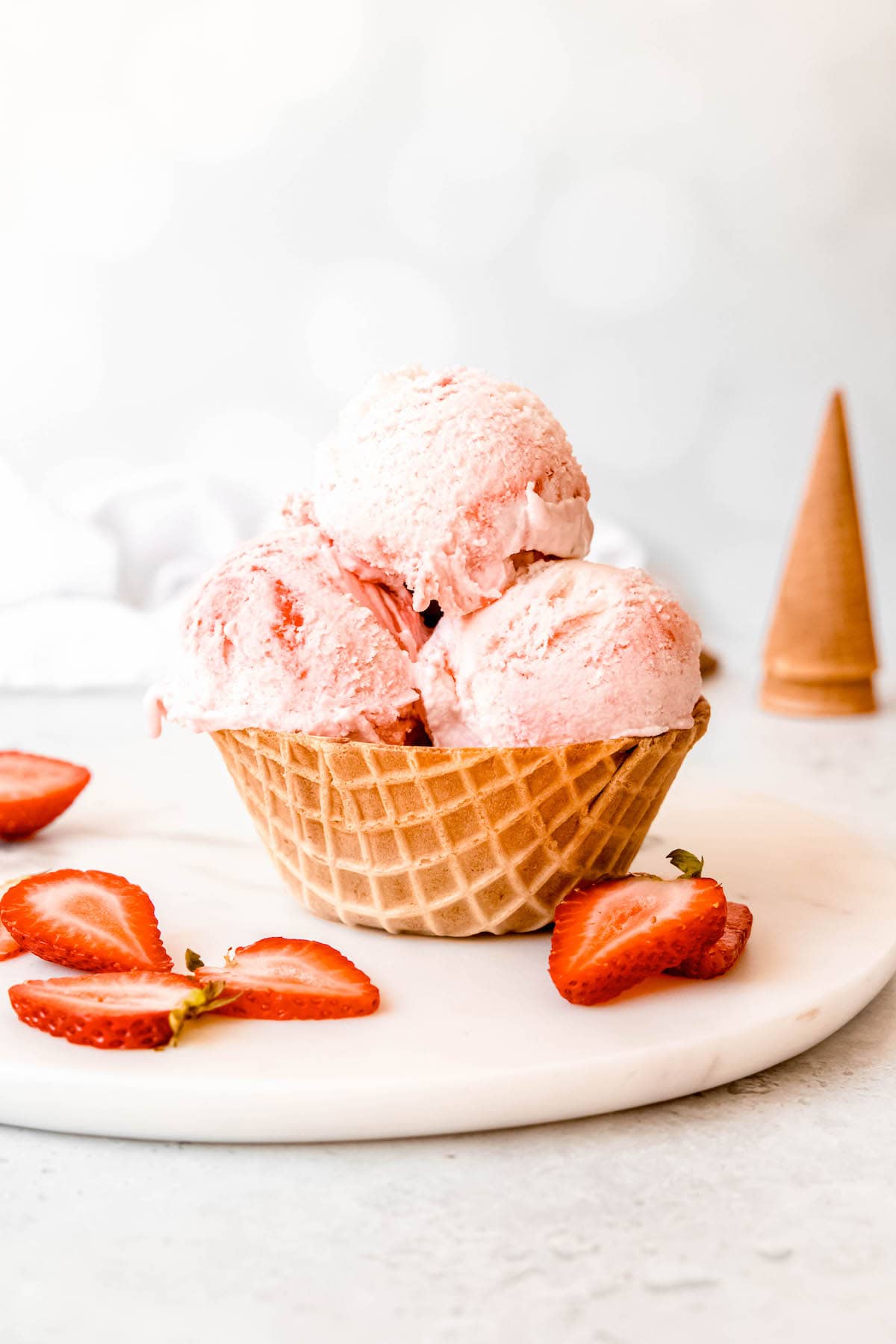 strawberry swirl ice cream cup on a white table with fresh sliced strawberries and a stack of ice cream cones in the background.