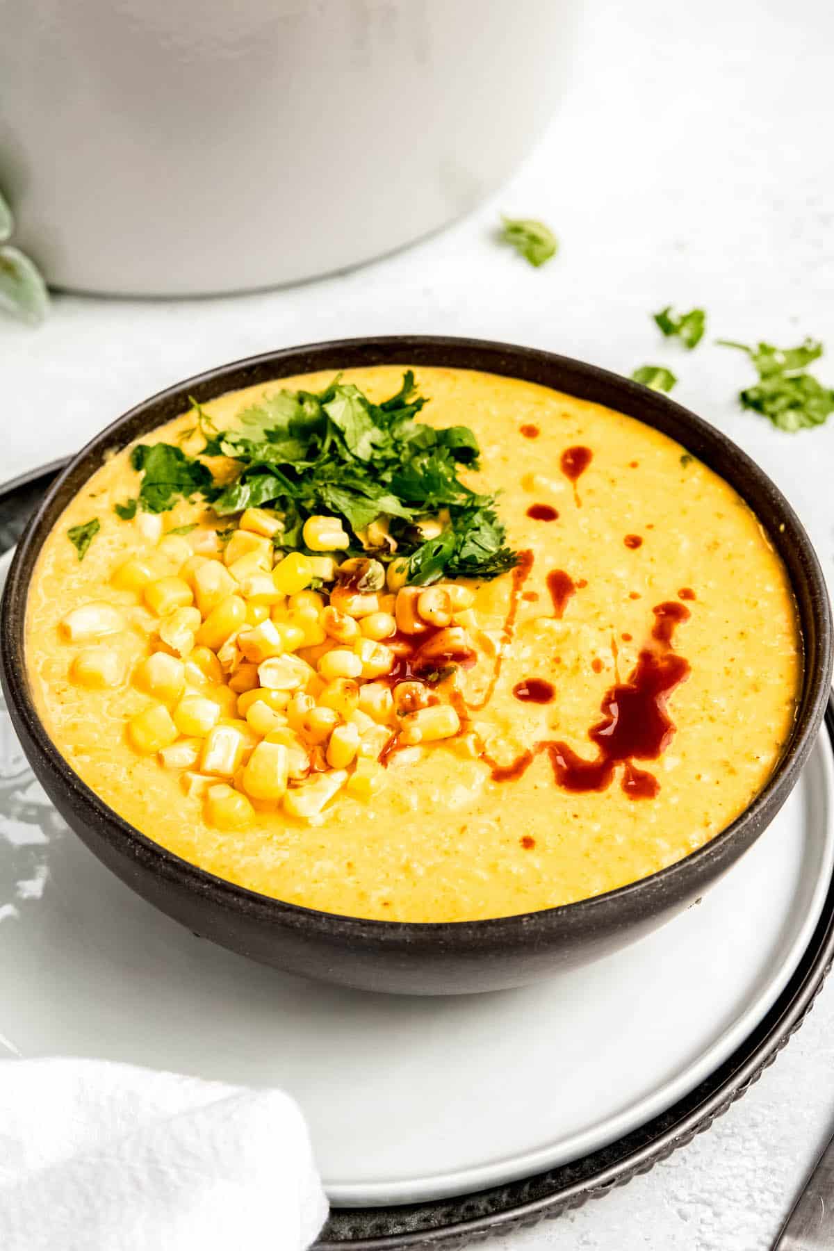 white dinner plate with a black bowl of bright yellow Panera-style mexican street corn chowder garnished with corn kernels, cilantro, and hot sauce in front of a white dutch oven.