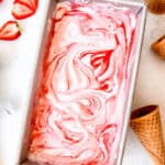 loaf tin filled with homemade strawberry swirl ice cream after freezing on a white table with ice cream cones and fresh strawberry slices.