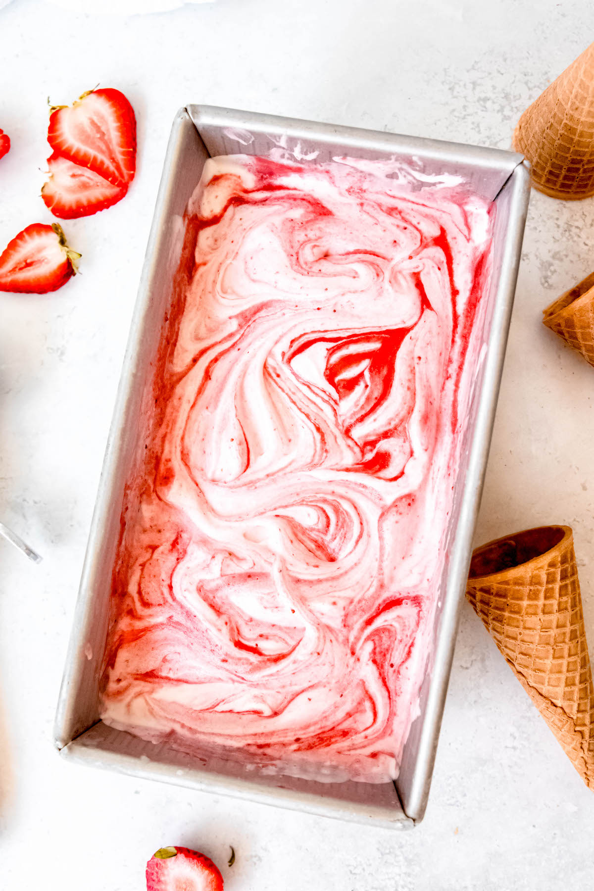 loaf tin filled with homemade strawberry swirl ice cream after freezing on a white table with ice cream cones and fresh strawberry slices.