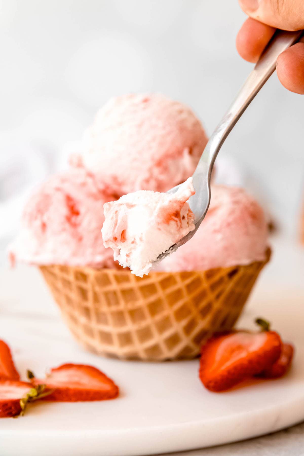spoonful of strawberry swirl ice cream in front of a waffle bowl filled with three scoops on a white table with fresh strawberry slices.