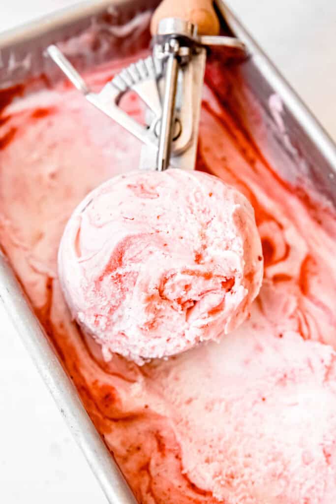45 degree angle shot of a spring-loaded ice cream scoop filled with vanilla strawberry swirl ice cream resting in the loaf pan.