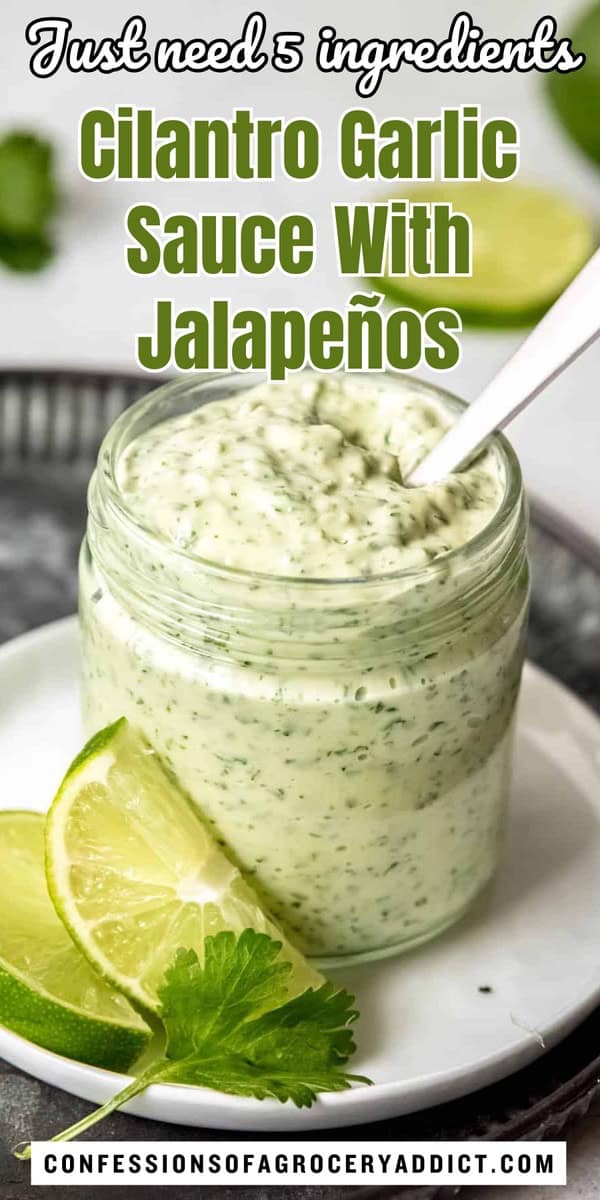 vertical pin with a photo of a jar of cilantro garlic lime sauce with a silver spoon sticking out and two lime slices and a sprig of cilantro in the foreground with text overlay that reads "just neeed 5 ingredients, cilantro garlic sauce with jalapeños."