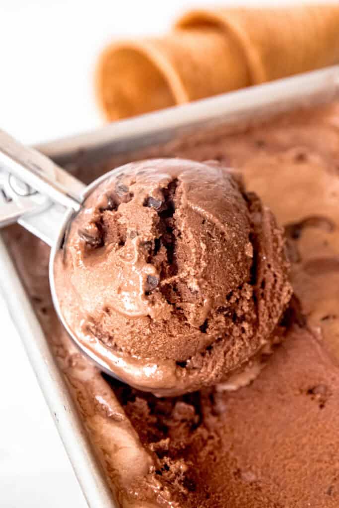 ice cream scoop filled with a scoop of chocolate chocolate chip ice cream resting in the loaf tin with the rest of the batch and a few waffle cones in the background.