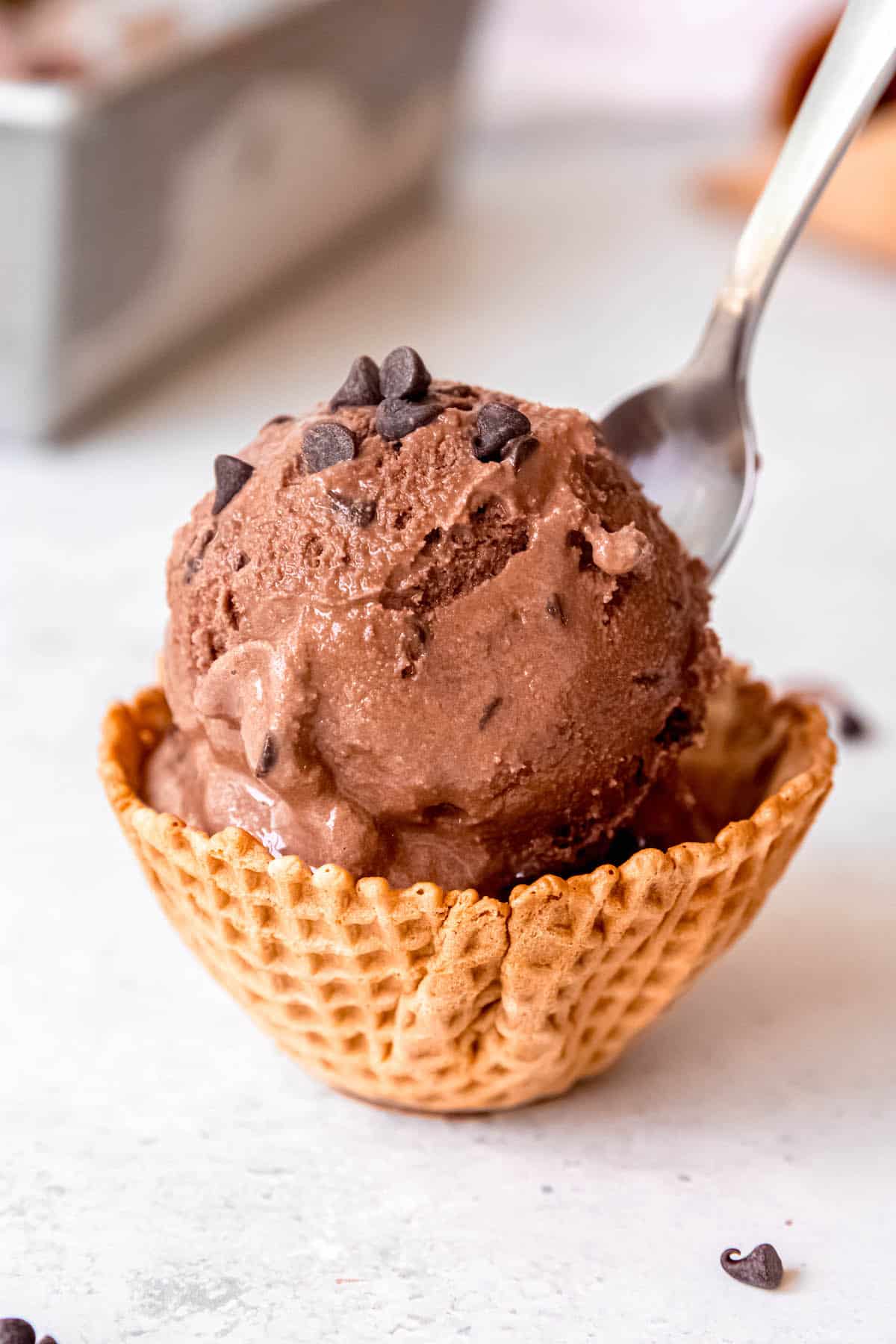 scoop of chocolate chocolate chip ice cream in a waffle cone bowl with a silver spoon sticking out and a few mini chocolate chips scattered on top and around.