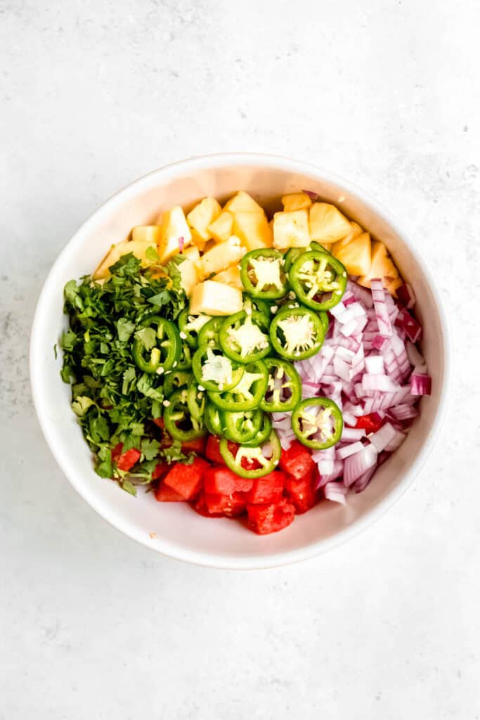 chopped pineapple, watermelon, jalapeno, red onion, and cilantro added to a large white salad bowl. 