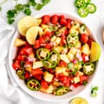 flat lay shot of a serving bowl filled with spicy watermelon salad with pineapple on a white serving table with fresh jalapeño slices, cilantro, and lime rounds scattered around.