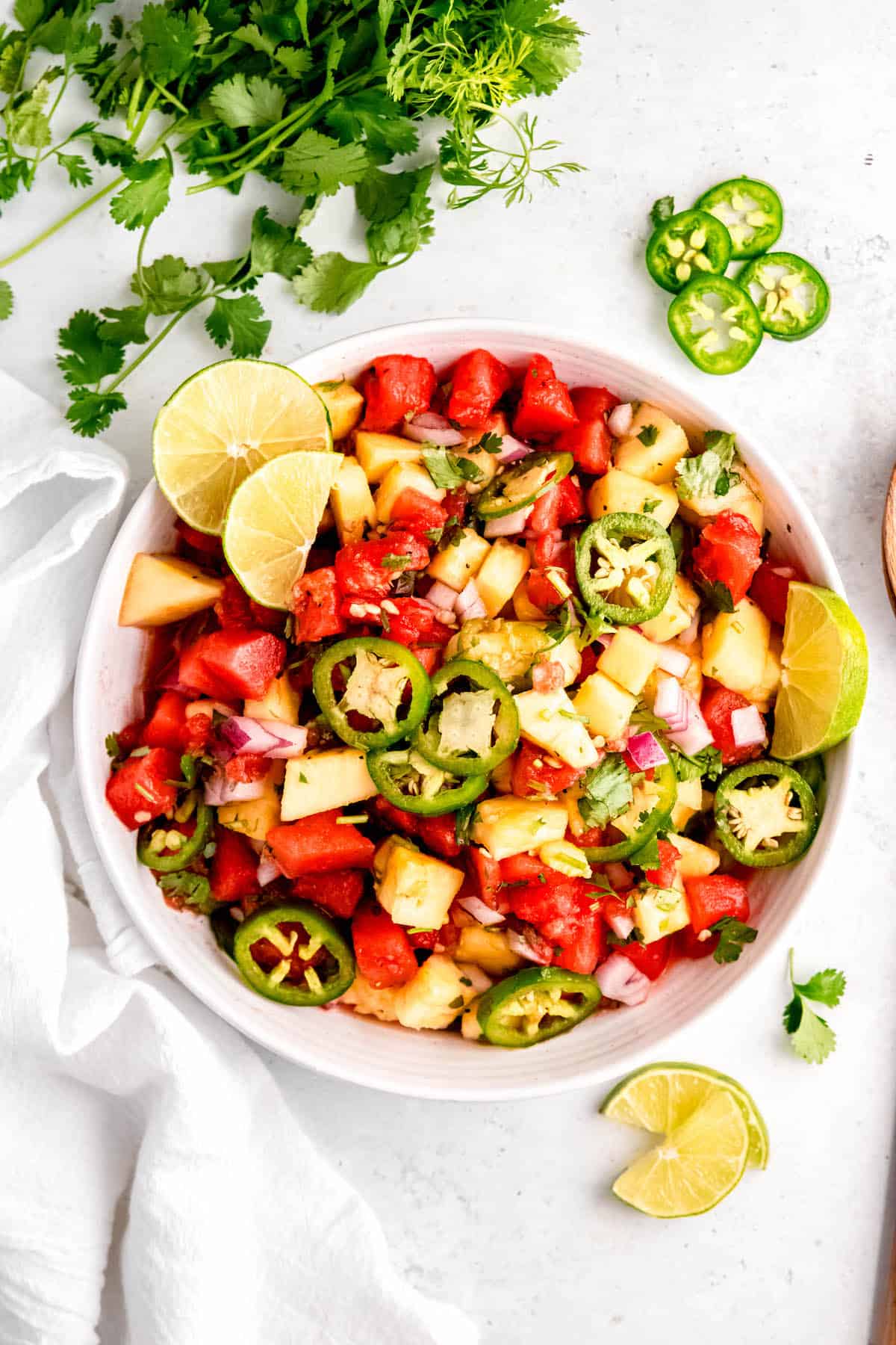 flat lay shot of a serving bowl filled with spicy watermelon salad with pineapple on a white serving table with fresh jalapeño slices, cilantro, and lime rounds scattered around.