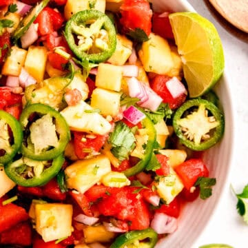 overhead shot of a round white serving bowl filled with spicy pineapple and watermelon salad on a white table with lime wedges and wooden salad claws.