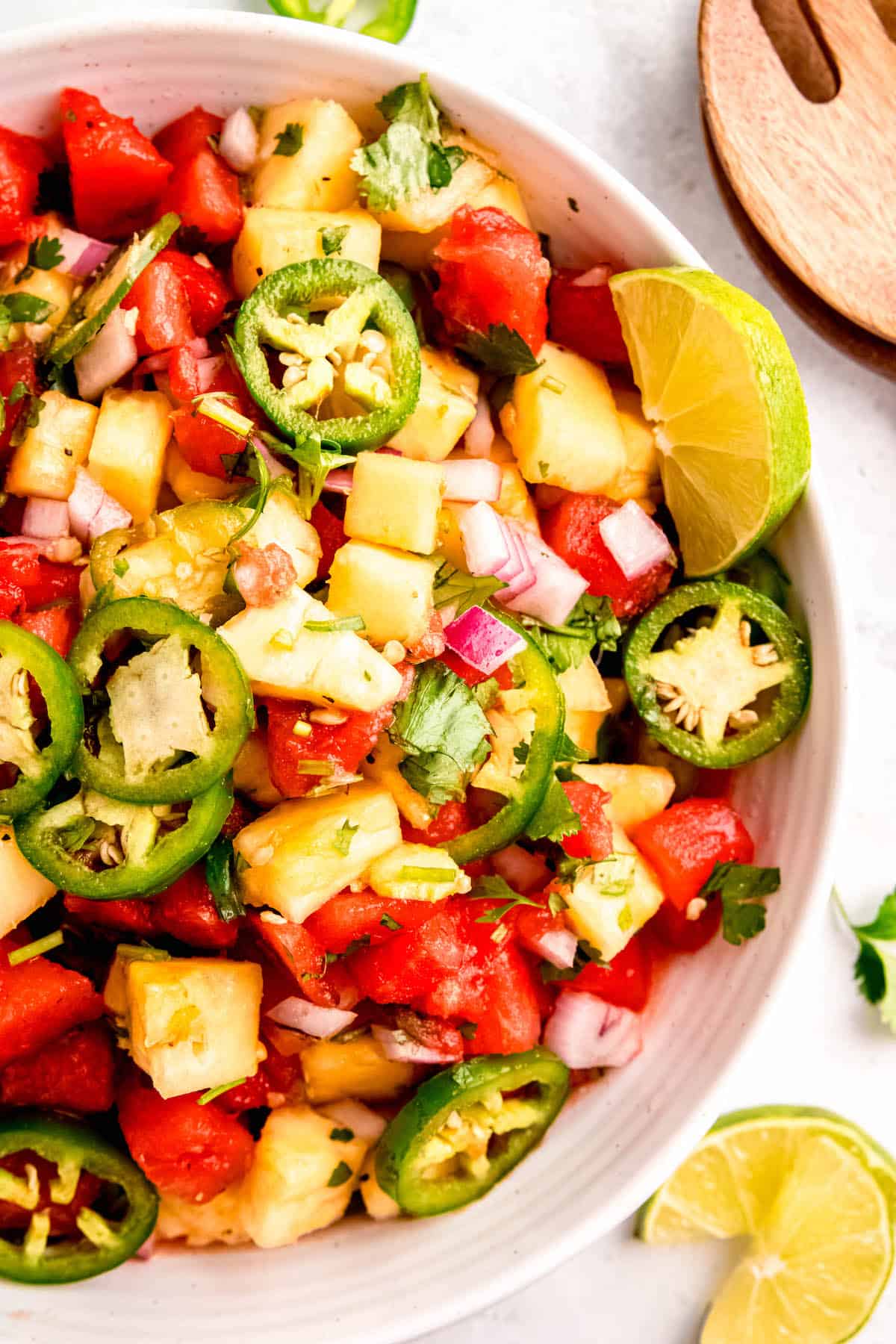 overhead shot of a round white serving bowl filled with spicy pineapple and watermelon salad on a white table with lime wedges and wooden salad claws.