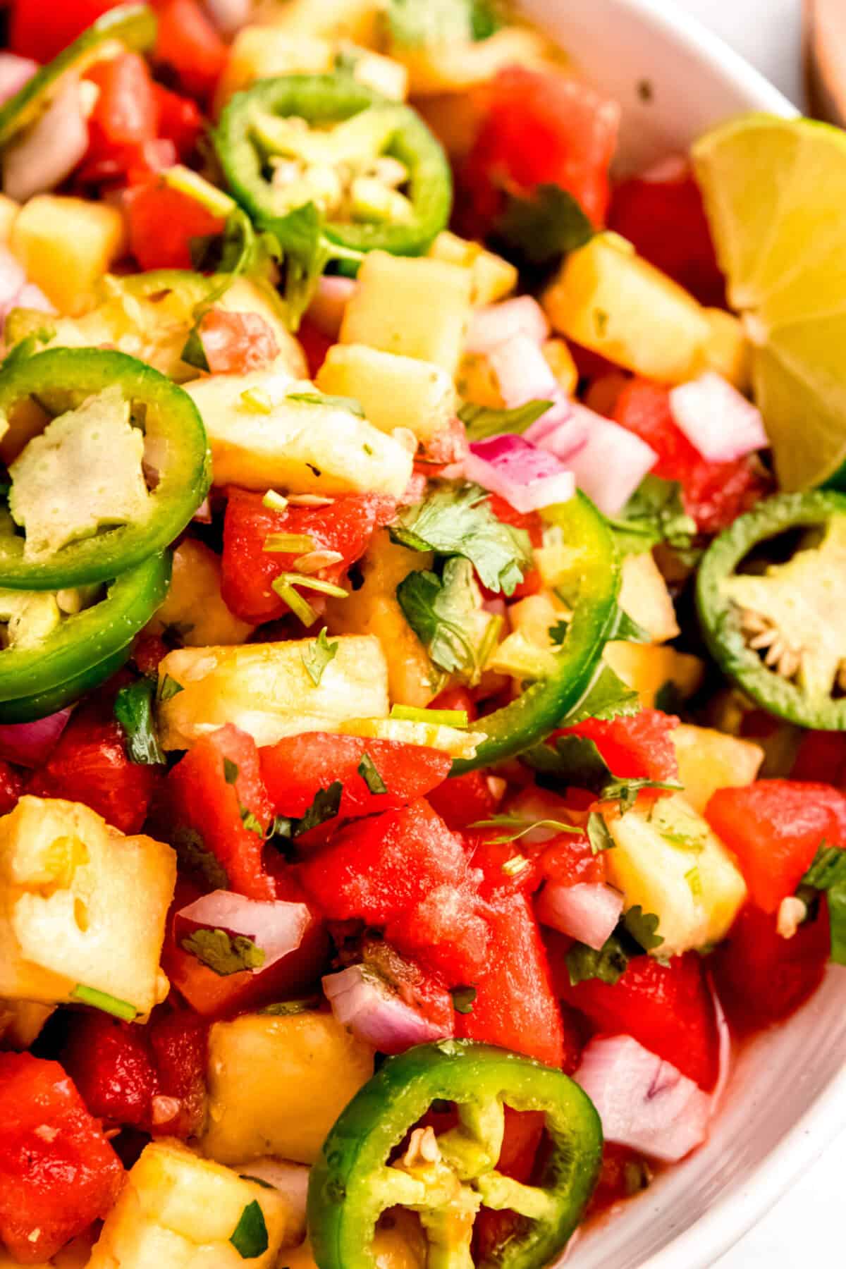 closeup overhead shot of spicy watermelon salad in a serving bowl showing the slices of jalapeños, cubes of pineapple, minced red onions, fresh cilantro, and a halved lime.