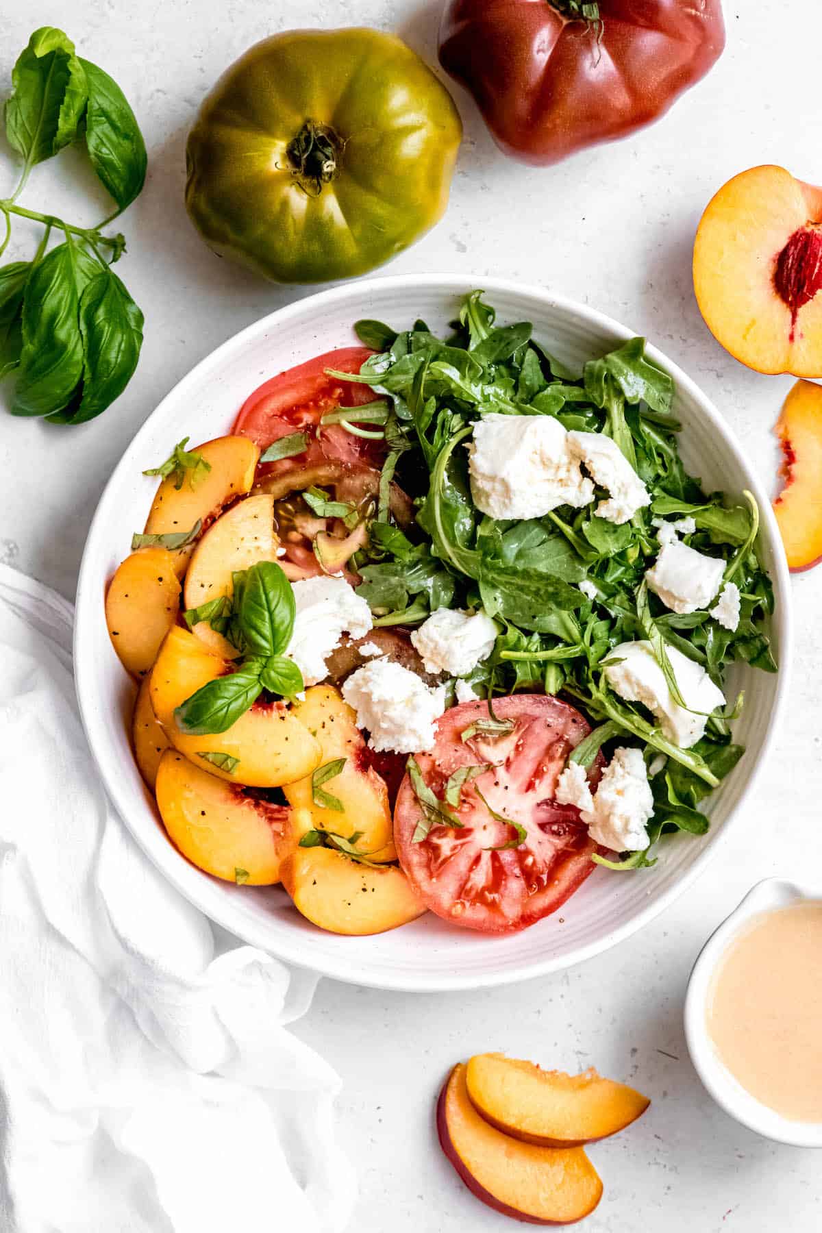 colorful arugula, tomato, burrata, and peach caprese salad in a white serving bowl with the whole ingredients scattered around the bowl.