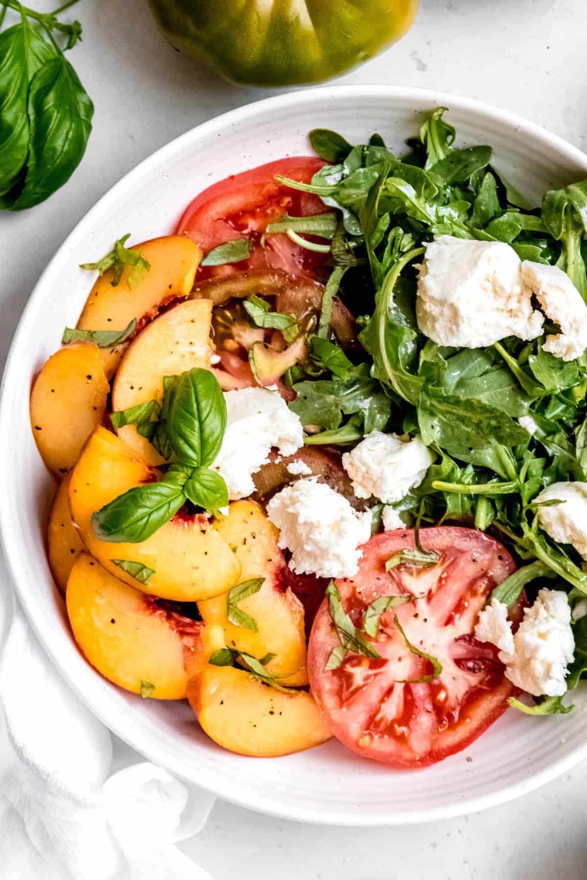 overhead shot of a bowl of burrata caprese with peaches, tomatoes, and white balsamic vinaigrette, with an heirloom green tomato and a sprig of fresh basil above.
