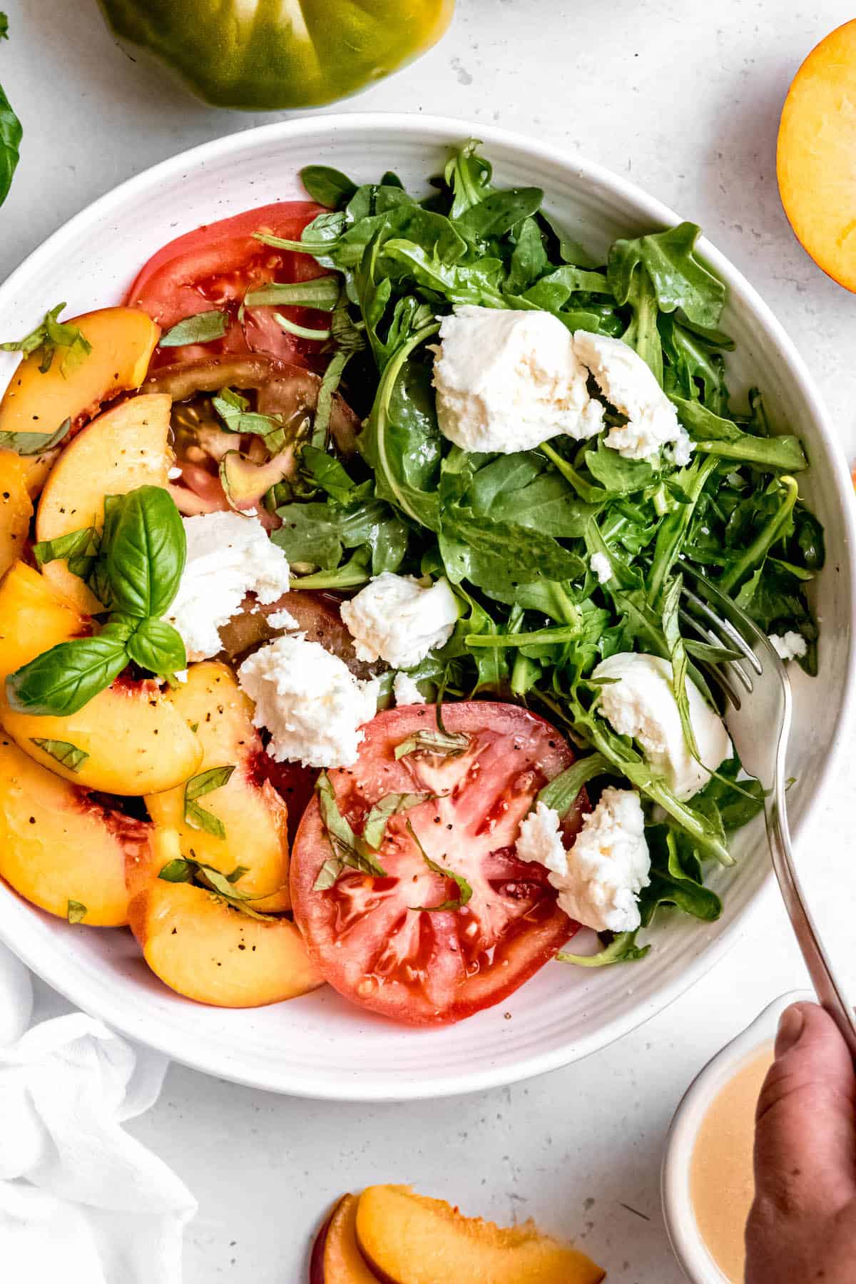 hand using a fork to take a bite of arugula and burrata from the tomato peach caprese salad bowl.