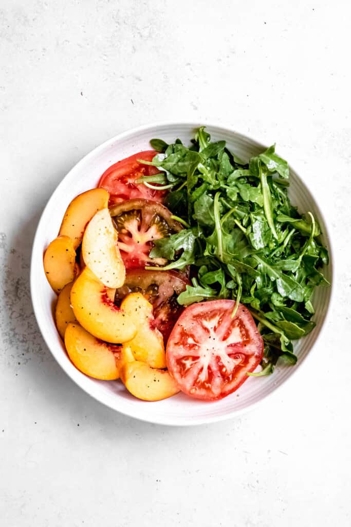dressed arugula, sliced tomatoes, and peaches in a white serving bowl.