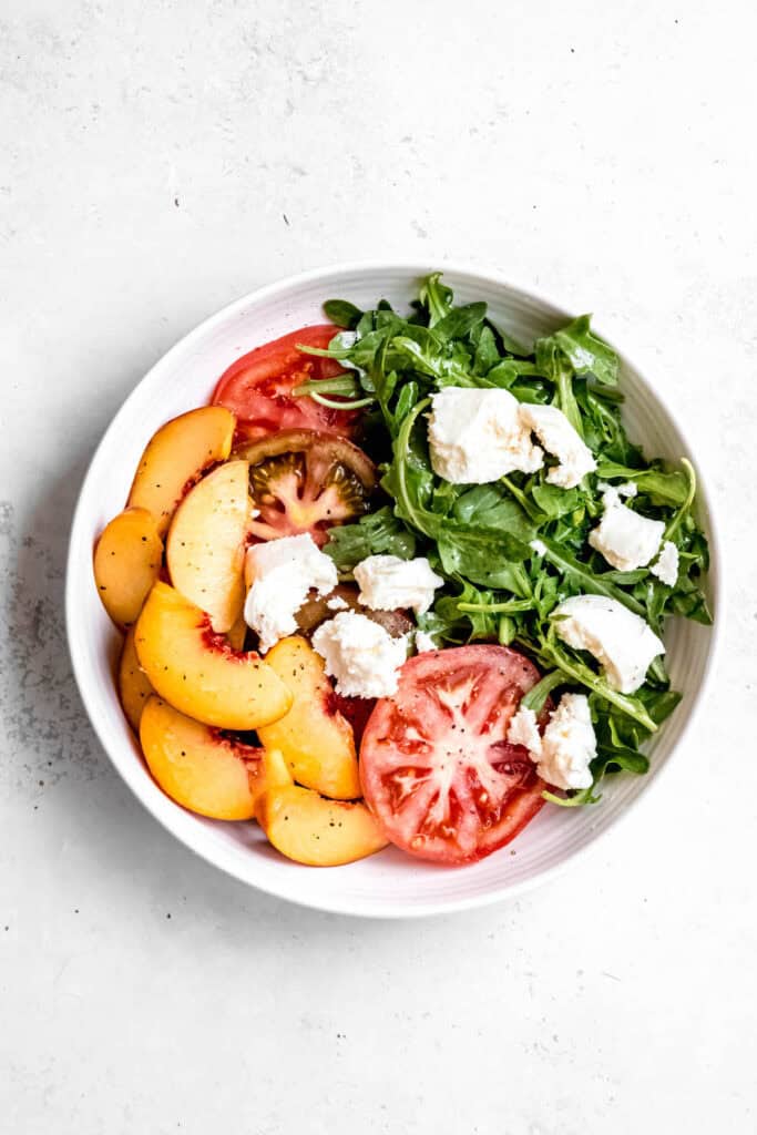 white serving bowl with peach burrata caprese after adding torn mozzarella and freshly cracked pepper.