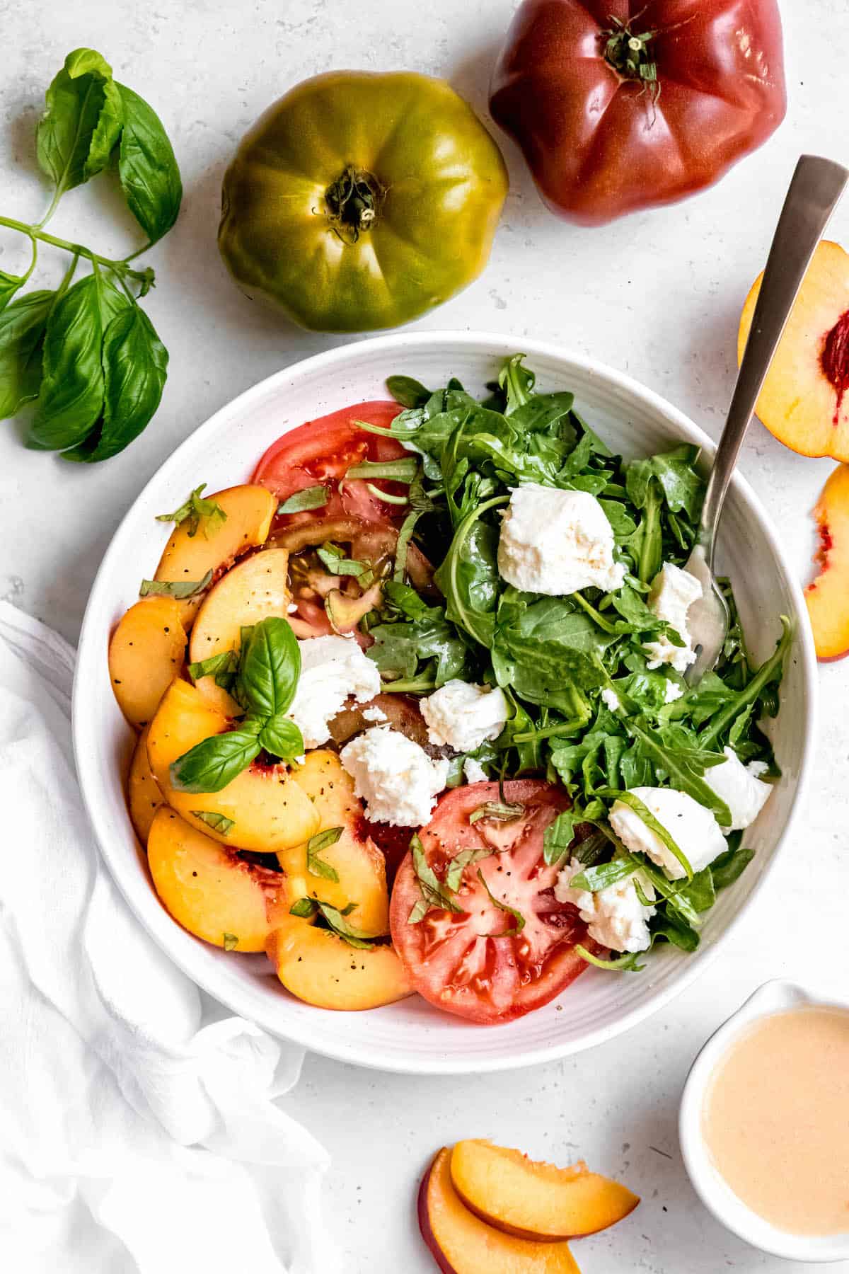 peach burrata salad as a fun twist on tomato caprese salad with arugula and white balsamic vinaigrette in a round white bowl with a silver fork, and fresh tomatoes, basil, and peaches scattered around.