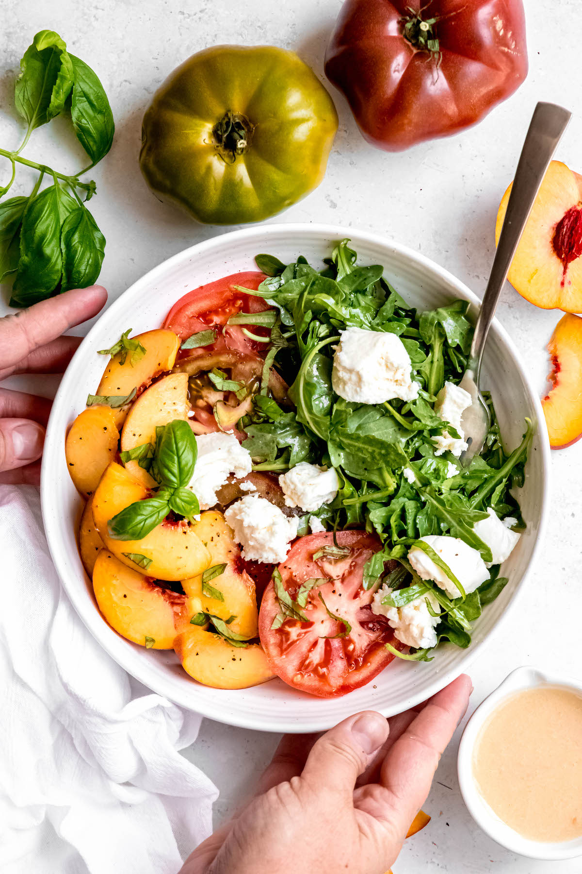 hands grabbing a round white bowl filled with burrata tomato peach caprese salad on a white table with heirloom tomatoes, fresh basil sprigs, sliced peaches, and a small carafe of homemade white balsamic vinaigrette.