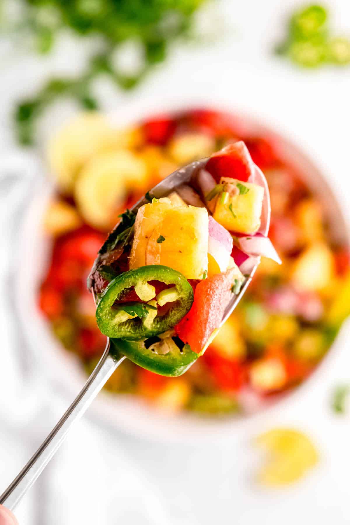 silver serving spoon with a scoop of spicy pineapple salad with watermelon, red onions, jalapeños, cilantro, and a tangy lime dressing with the bowl of the salad on a white table with a bunch of cilantro blurred out below.