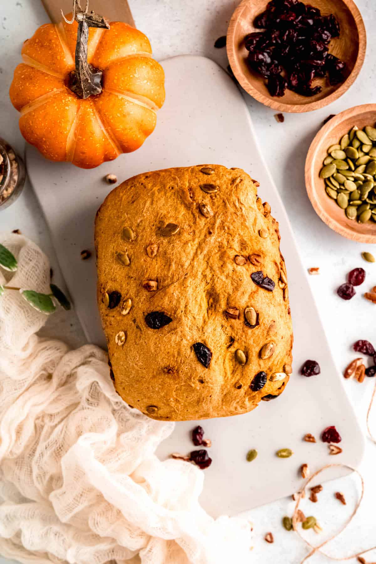 overhead shot of a loaf of pumpkin bread machine bread on a white cutting board with dried cranberries, pepitas, and walnuts scattered around and pressed into the top of the loaf.