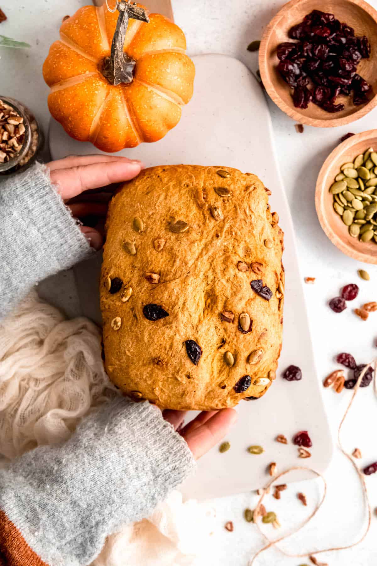 sweatered hands grabbing a freshly baked loaf of pumpkin bread machine bread from a white cutting board surrounded by wooden bowls of nuts, seeds, and dried fruit.