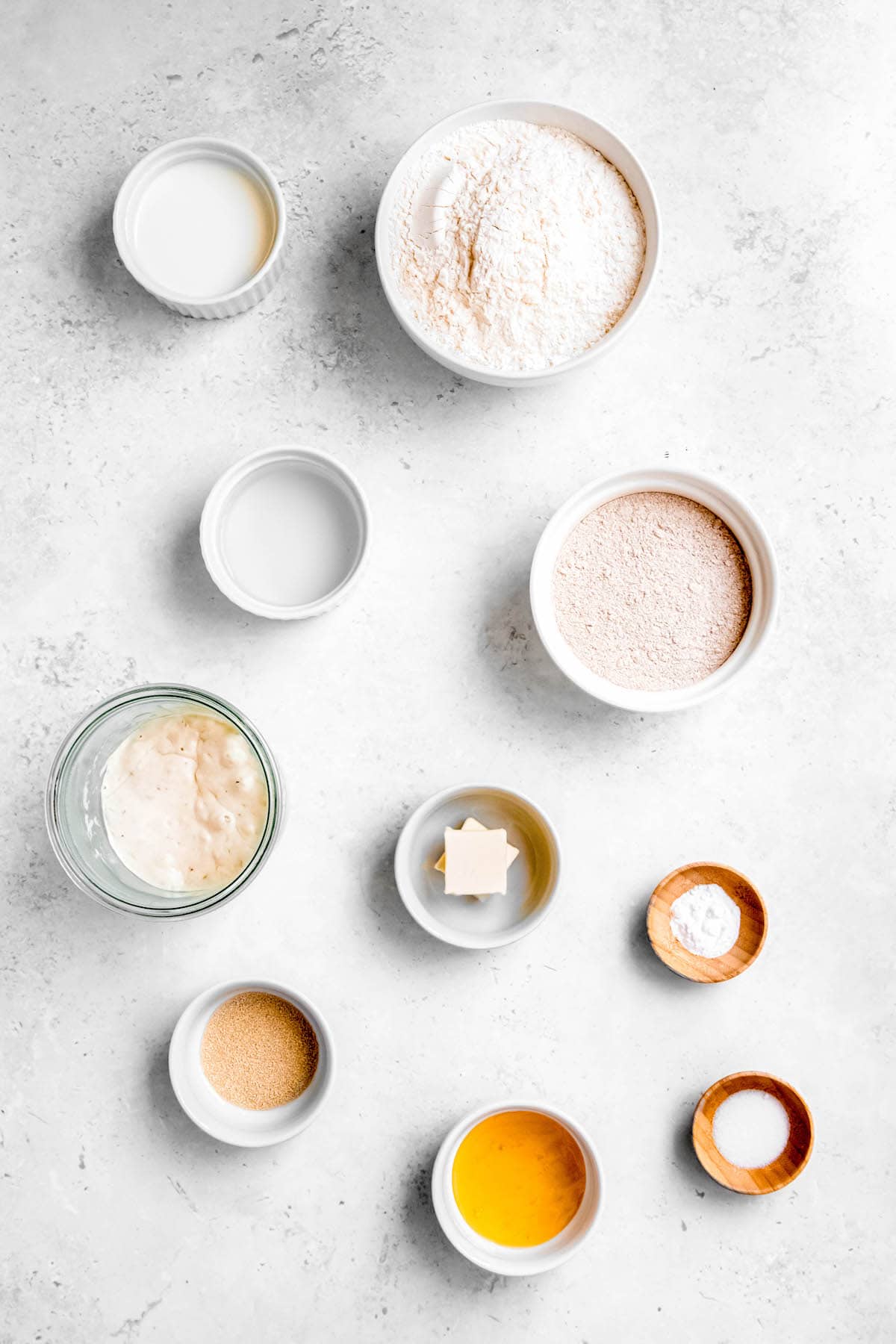 overhead shot of the ingredients to make sourdough discard sandwich bread in the bread machine all measured out in bowls on a white table.