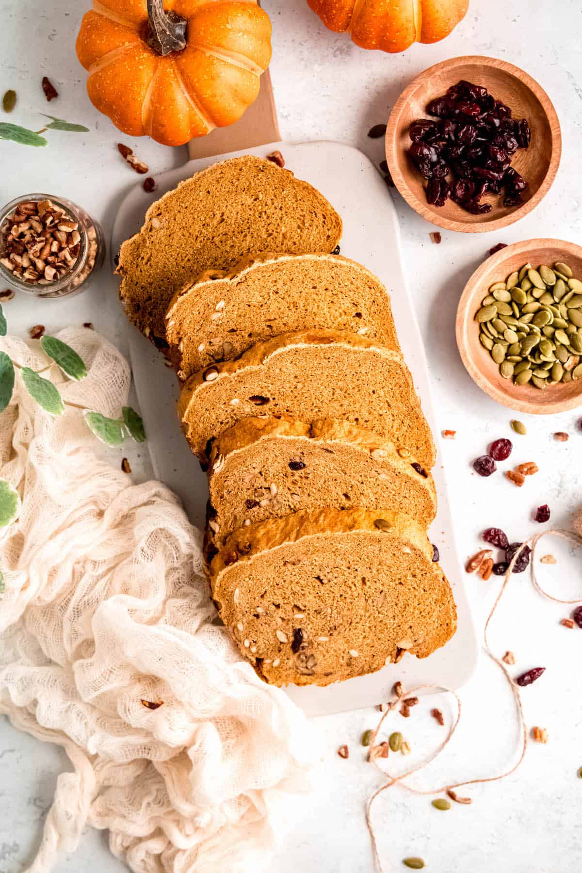 sliced loaf of pumpkin spice bread machine bread on aa white table with two mini pumpkins and wooden bowls of walnuts, pumpkin seeds, and dried cranberries.