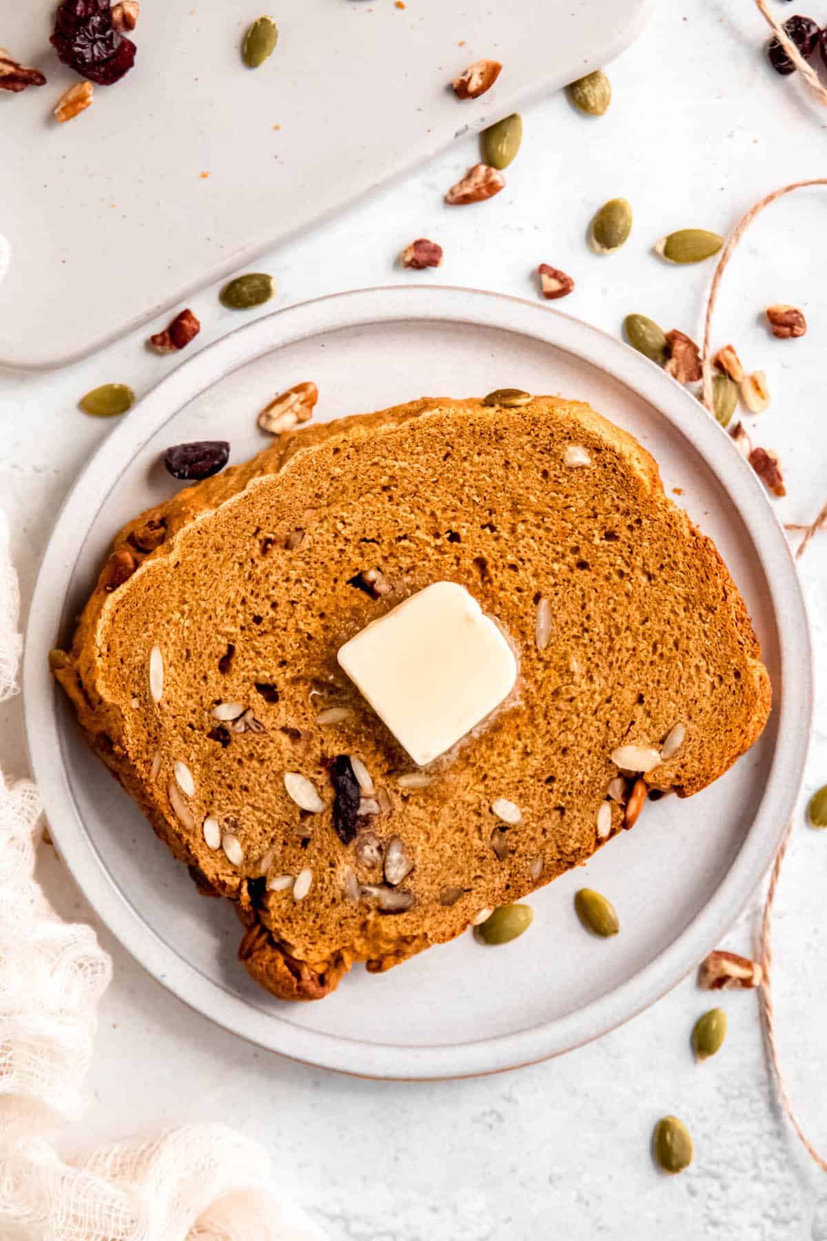 slice of pumpkin bread machine bread topped with a pat of butter on a white plate with nuts, seeds, and dried cranberries scattered around.