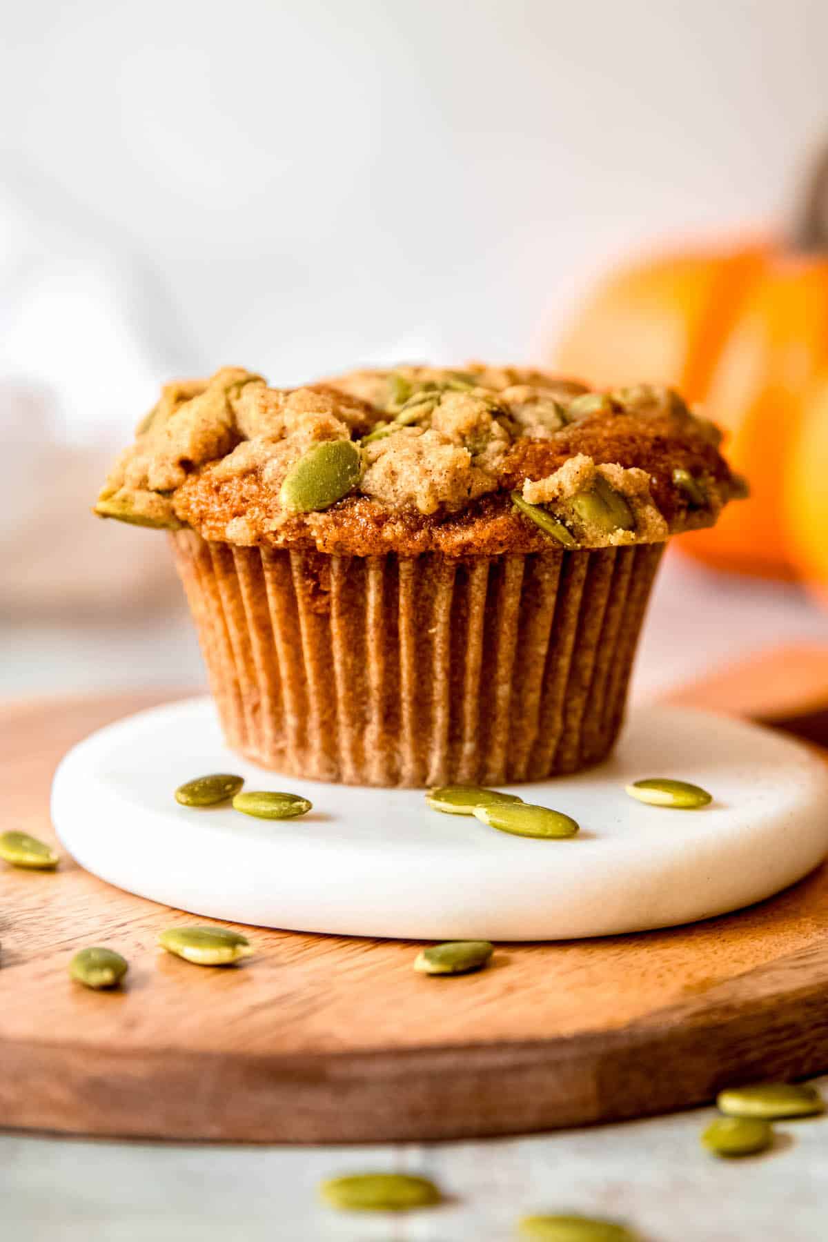 side on view of a banana pumpkin muffin topped with streusel on a white coaster on top of a wooden serving tray with orange pumpkins blurred in the background and hulled pumpkin seeds in the foreground.