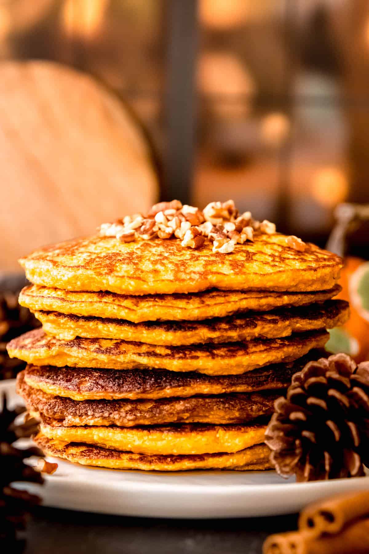 side on shot of a stack of oat protein pumpkin pancakes in front of a lit window and topped with chopped pecans.