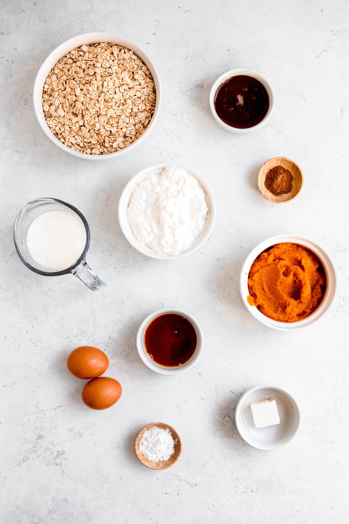 flat lay shot of the ingredients needed to make oatmeal cottage cheese pumpkin protein pancakes measured out into bowls on a white table.