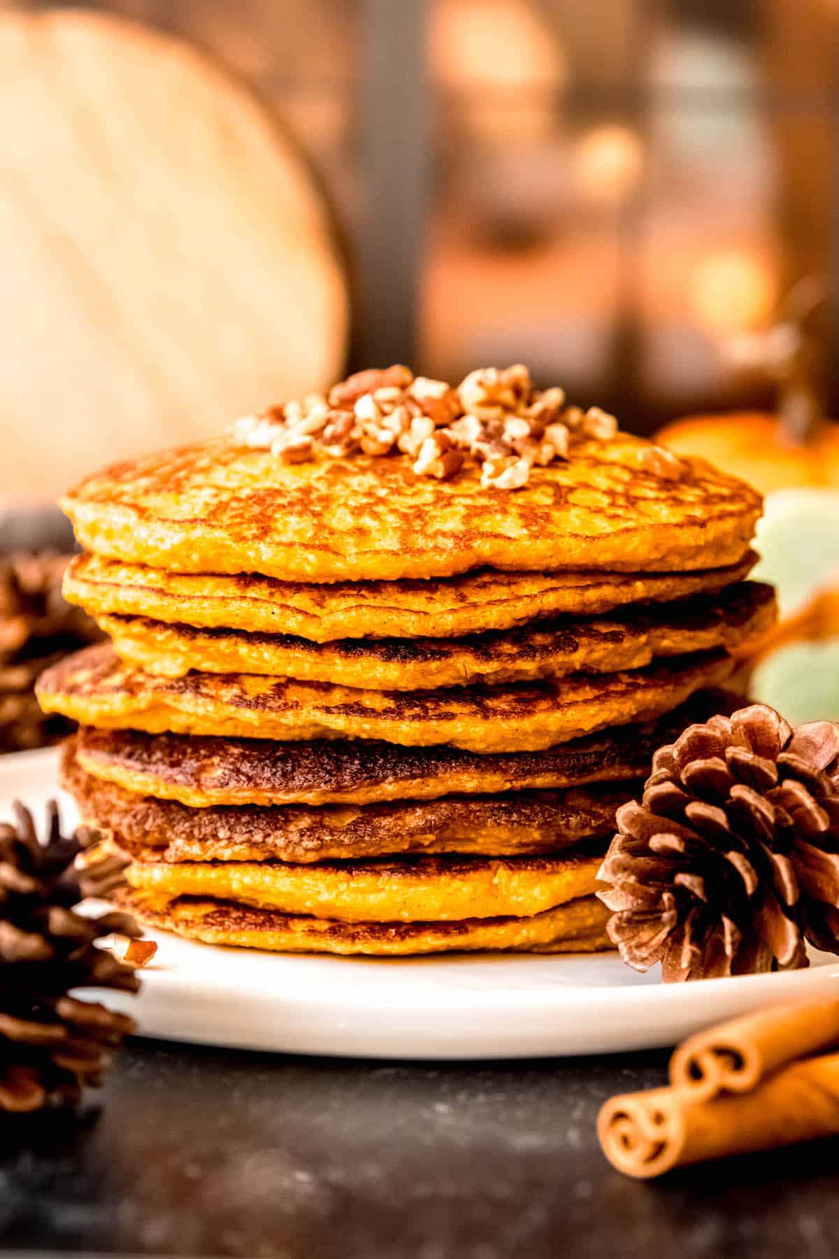 side on shot of a tall stack of protein-packed oatmeal cottage cheese pumpkin pancakes on a table with pine cones and cinnamon sticks and a window lit by fairy lights in. the background.