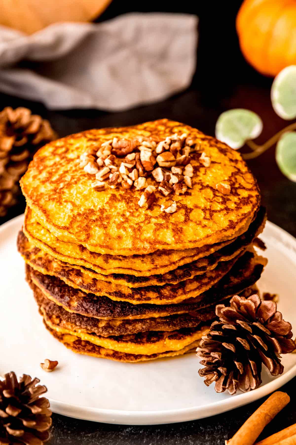 45 degree angle of a stack of golden brown pumpkin protein pancakes on a round white plate atop a black table with mini pumpkins, a green houseplant, pinecones, and cinnamon sticks.